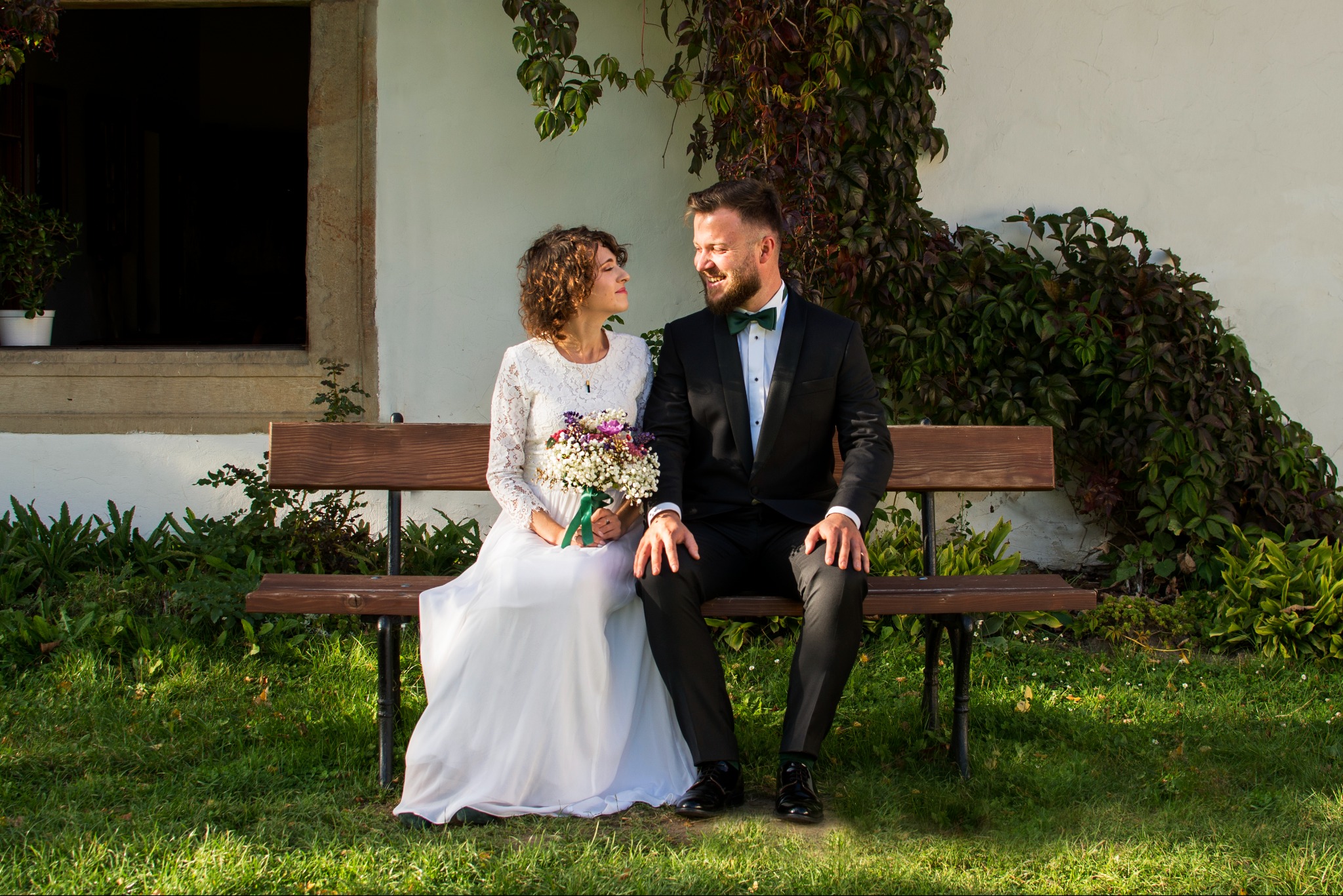 Para młoda siedzi na ławce przed budynkiem porośniętym bluszczem. Ona w koronkowej sukni, on w smokingu z zieloną muchą, patrzą na siebie z uśmiechem. Romantyczna sesja ślubna.