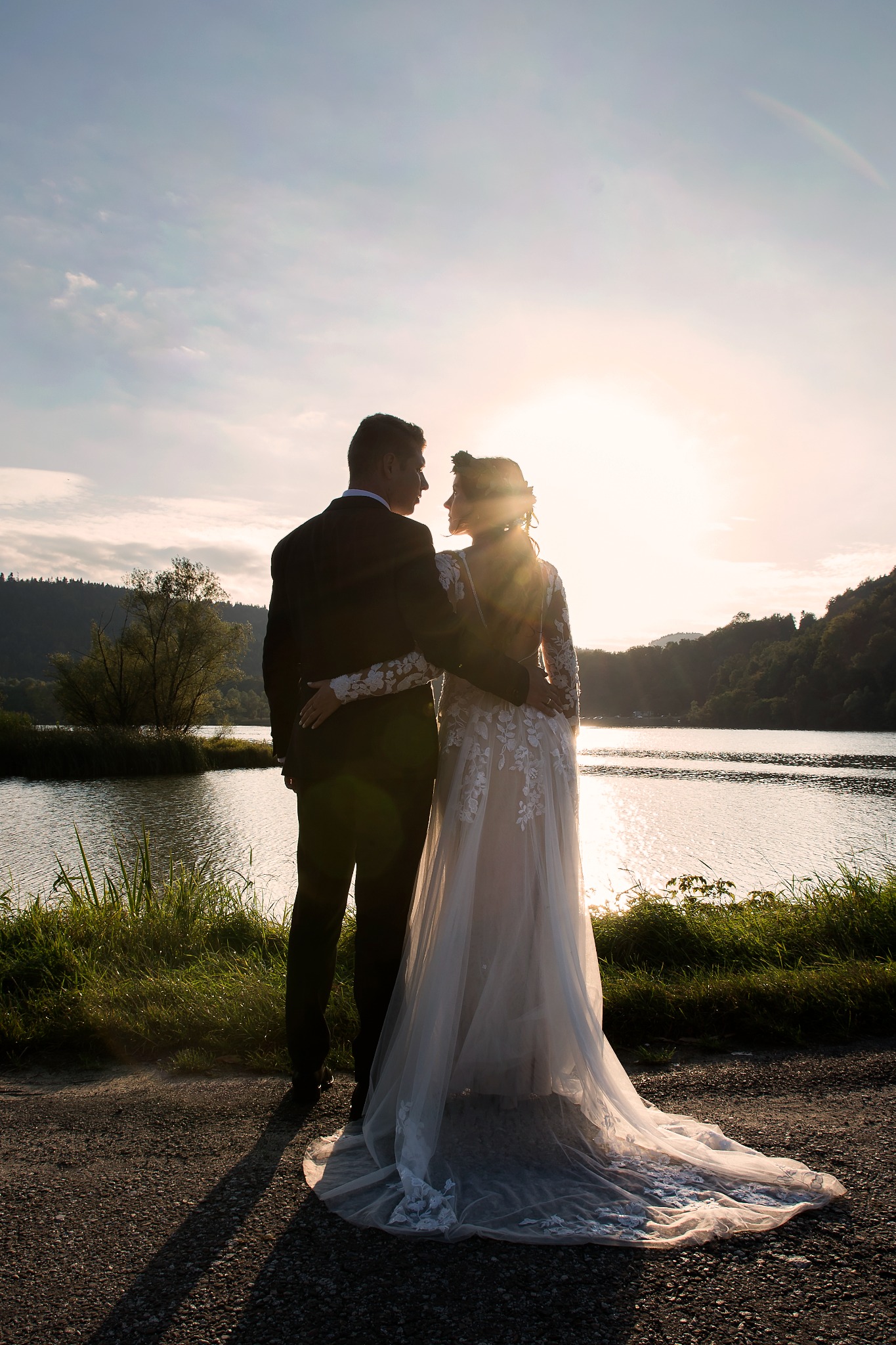 Para młoda, w kontraście do zachodzącego słońca nad jeziorem, obejmuje się. Suknia ślubna z koronkowymi detalami. Romantyczna sceneria zalesionych brzegów.