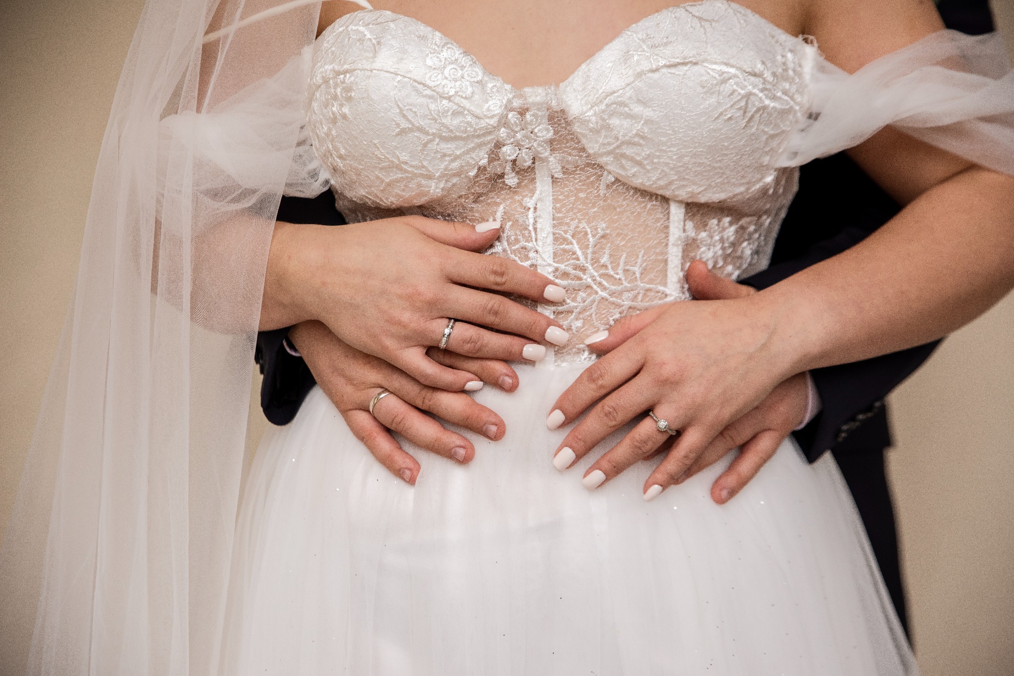 Zbliżenie na parę młodą: dłonie z obrączkami spoczywają na koronkowej sukni ślubnej. W tle fragment welonu i ciemny garnitur, ciepłe oświetlenie.