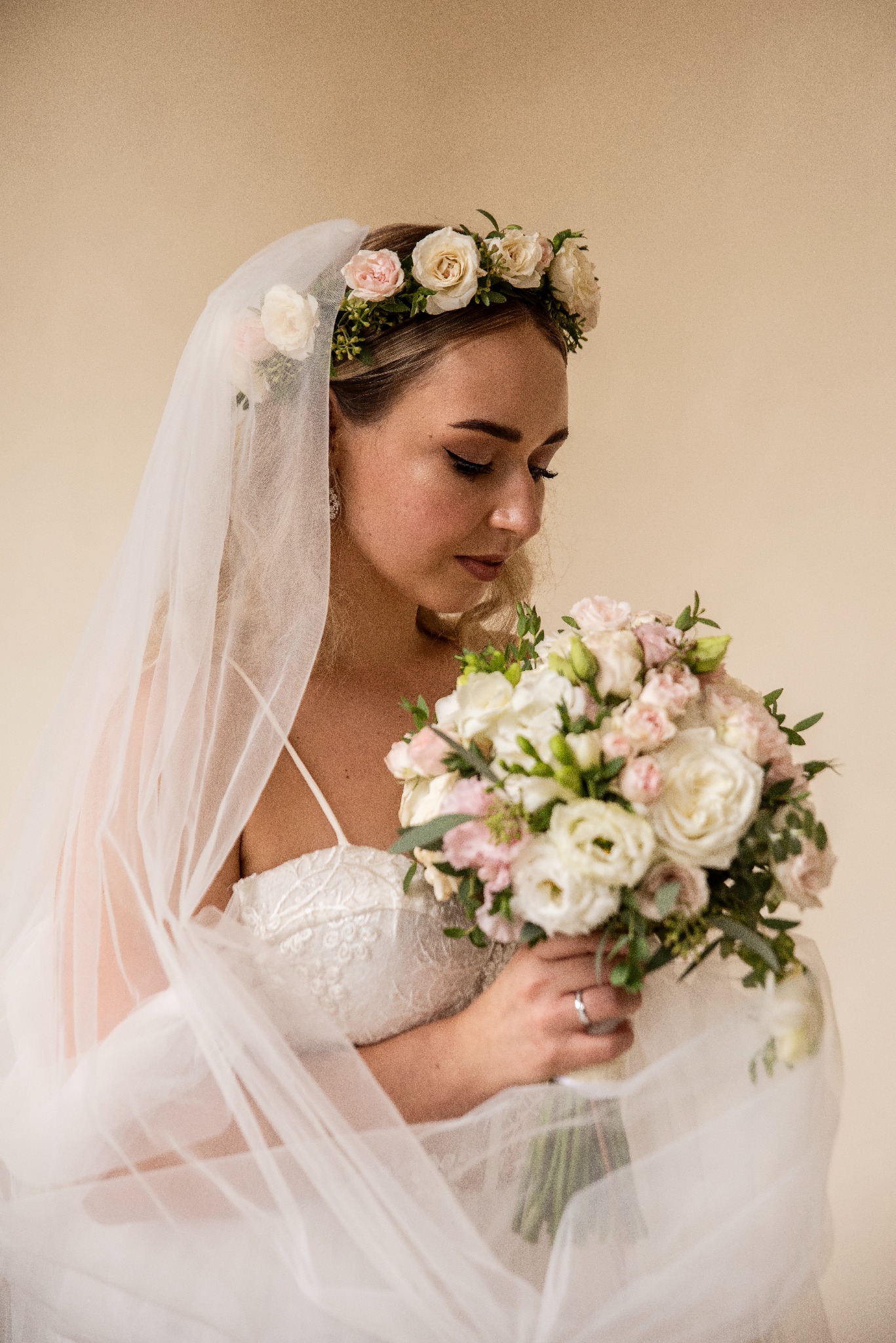 Panna młoda w białej sukni z welonem i wiankiem na głowie, trzyma bukiet ślubny, wzrok skierowany w dół. Delikatne oświetlenie i jasne tło.