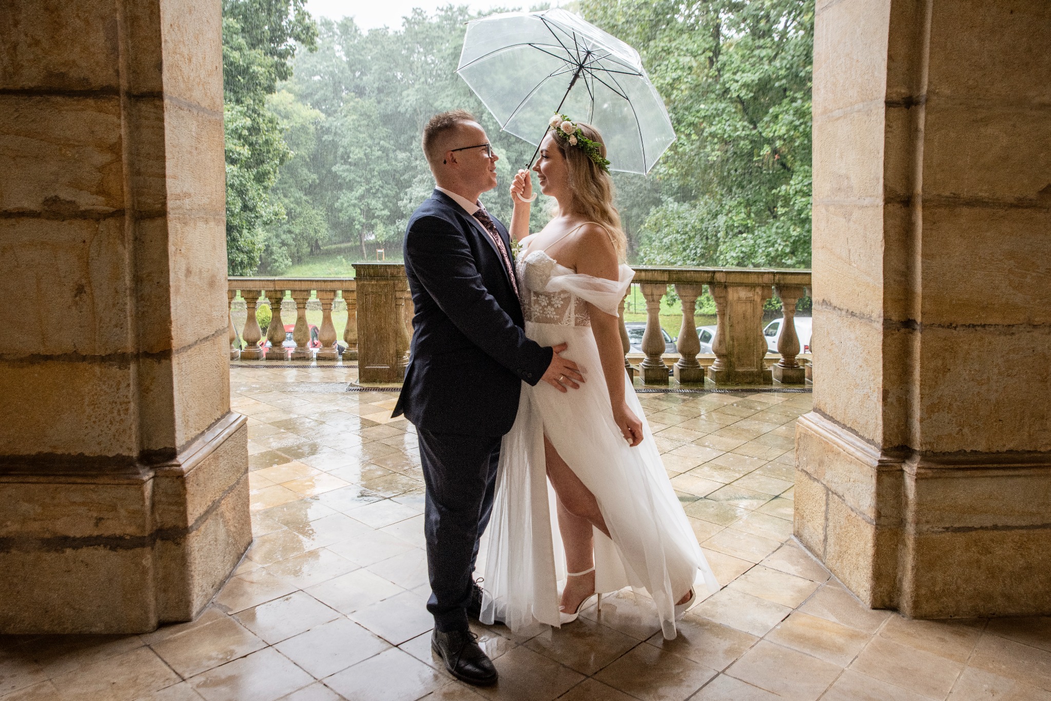 Para młoda pod przezroczystym parasolem w deszczowy dzień, stojąca na balkonie z kamiennymi kolumnami, w tle zieleń drzew.