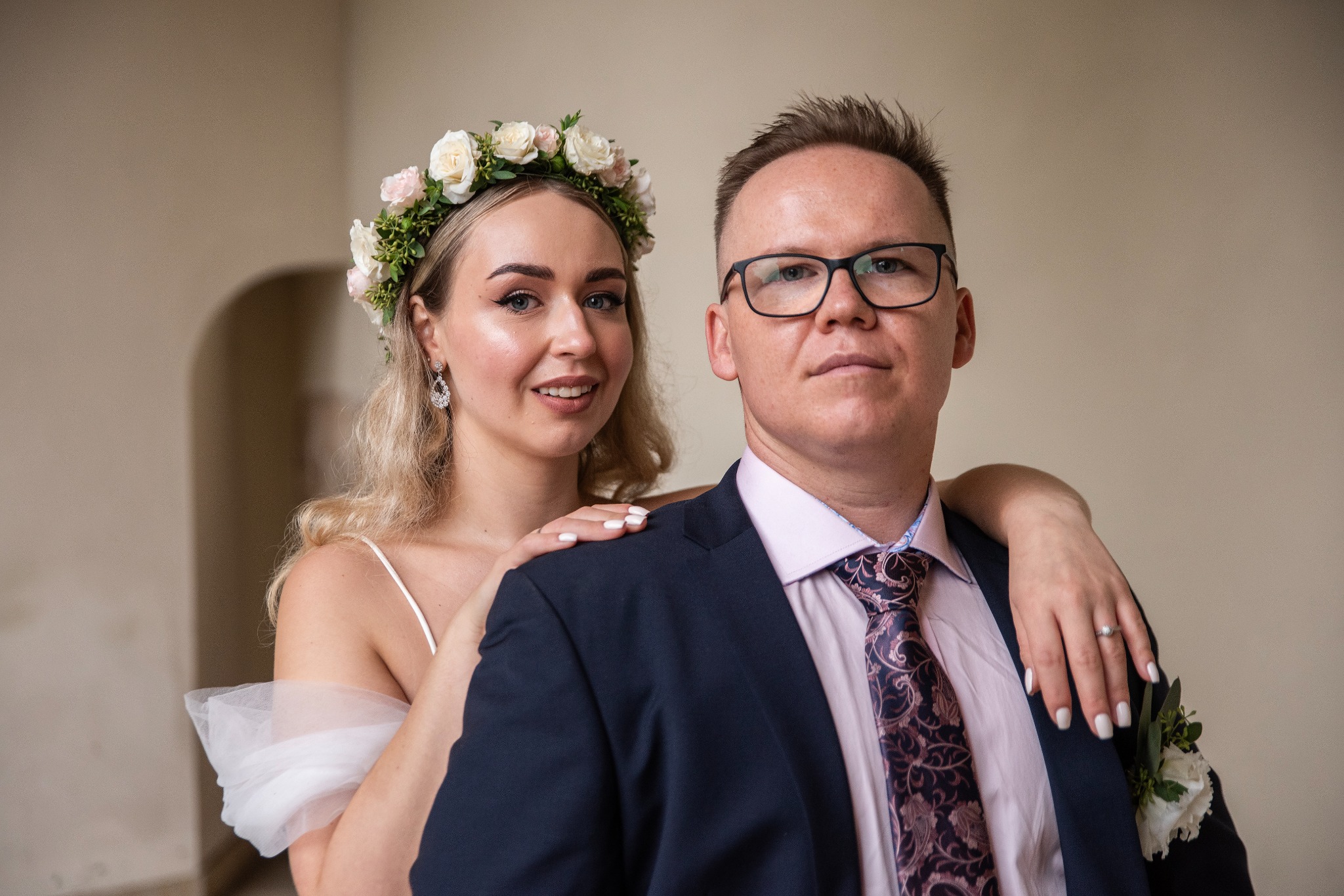 Para w strojach ślubnych: blondynka z wiankiem i mężczyzna w garniturze. Ona opiera ręce na jego ramionach, oboje uśmiechnięci, neutralne tło.