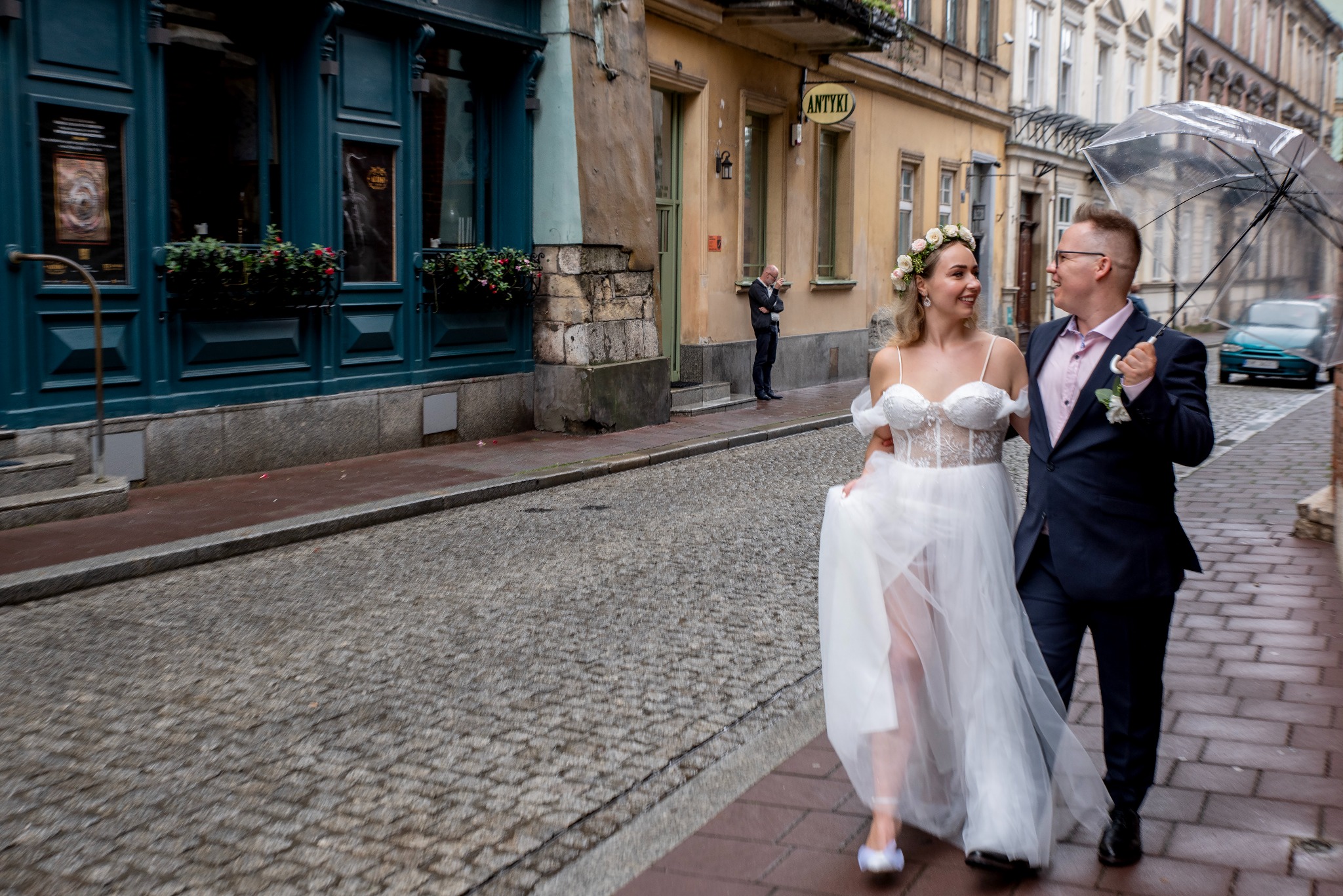 Uśmiechnięta para młoda spaceruje ulicą Starego Miasta pod przezroczystym parasolem. W tle budynki z szyldem 'Antyki' i mężczyzna w garniturze.