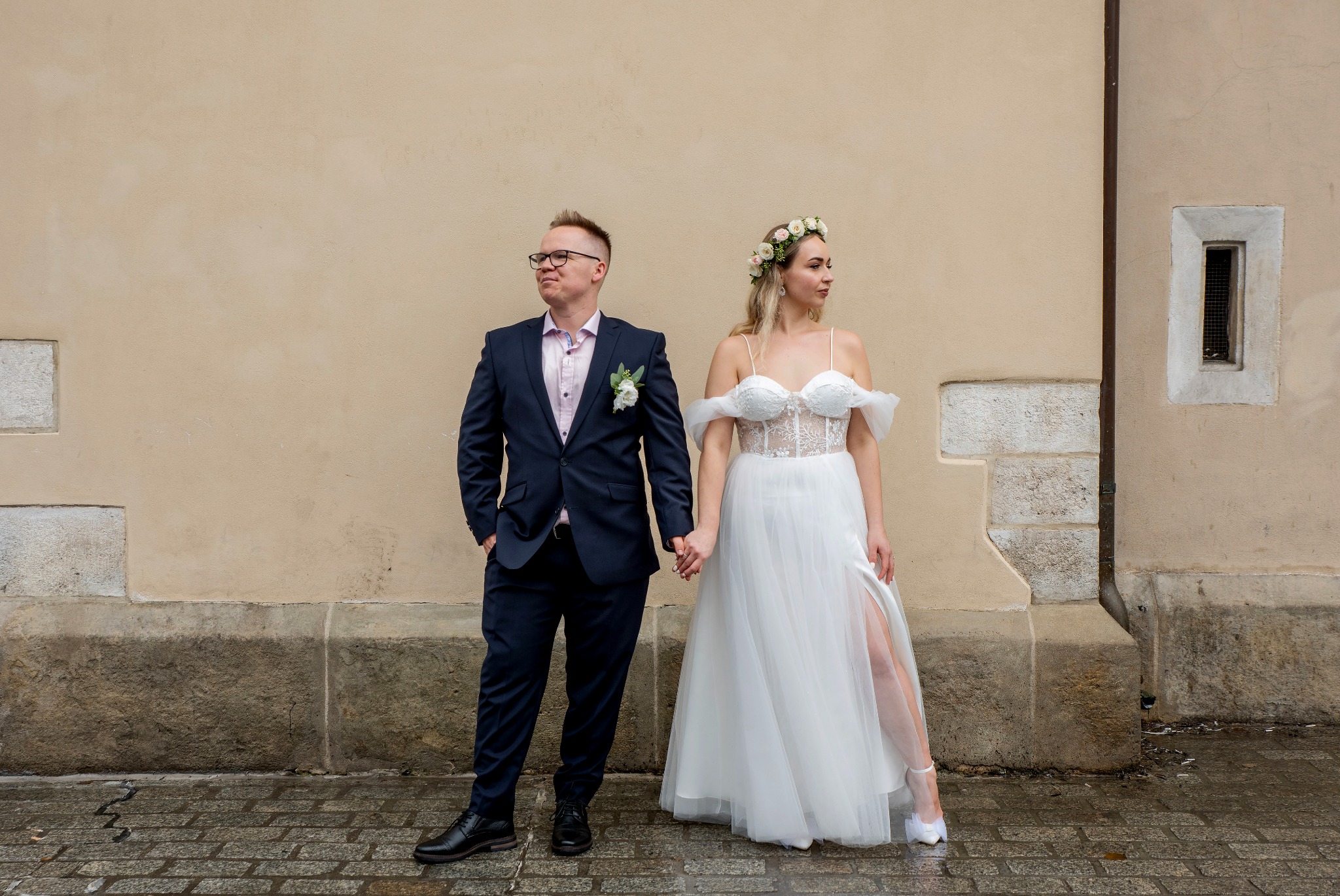 Para młoda trzymająca się za ręce na tle beżowej ściany z elementami architektonicznymi. Panna młoda w białej sukni z wiankiem, pan młody w granatowym garniturze.
