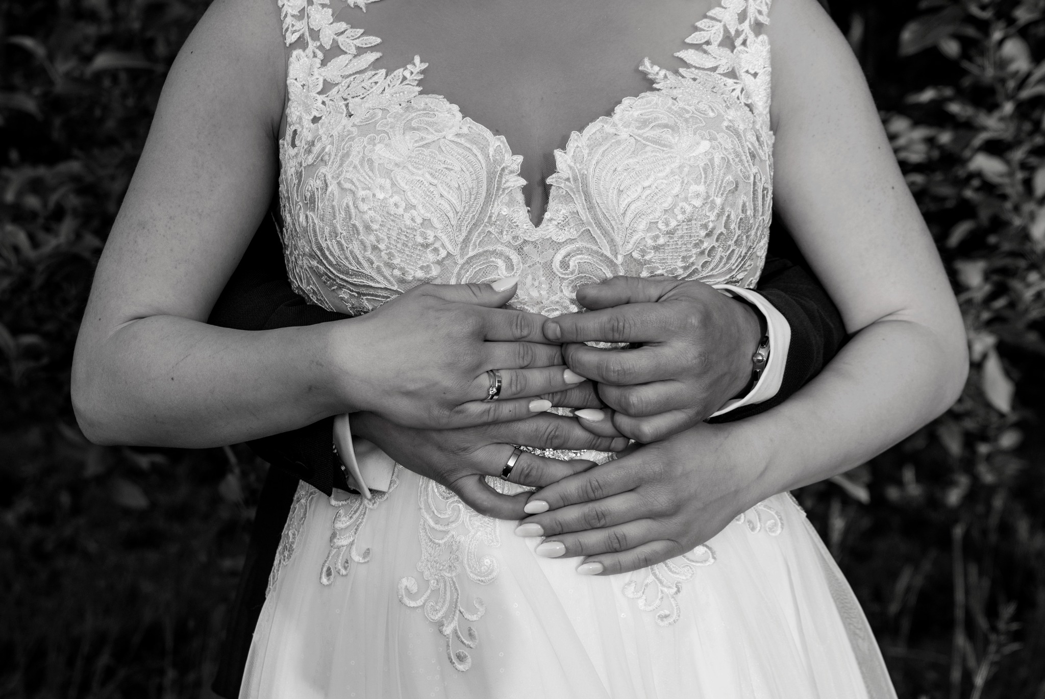 Czarno-białe zbliżenie na parę młodą: dłonie mężczyzny obejmują talię kobiety w koronkowej sukni ślubnej, eksponując obrączki na ich palcach.