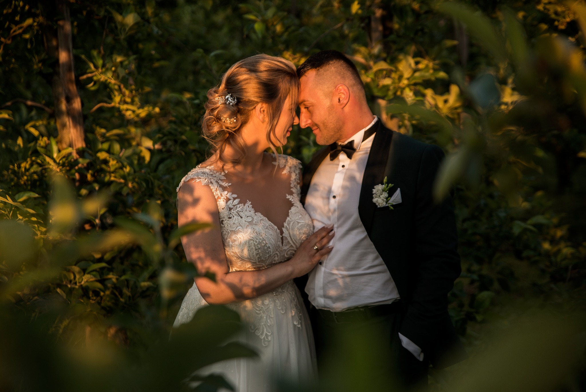 Para młoda w objęciach, delikatne światło podkreśla koronkową suknię panny młodej i bukiet pana młodego, tło z zieleni tworzy intymną atmosferę.