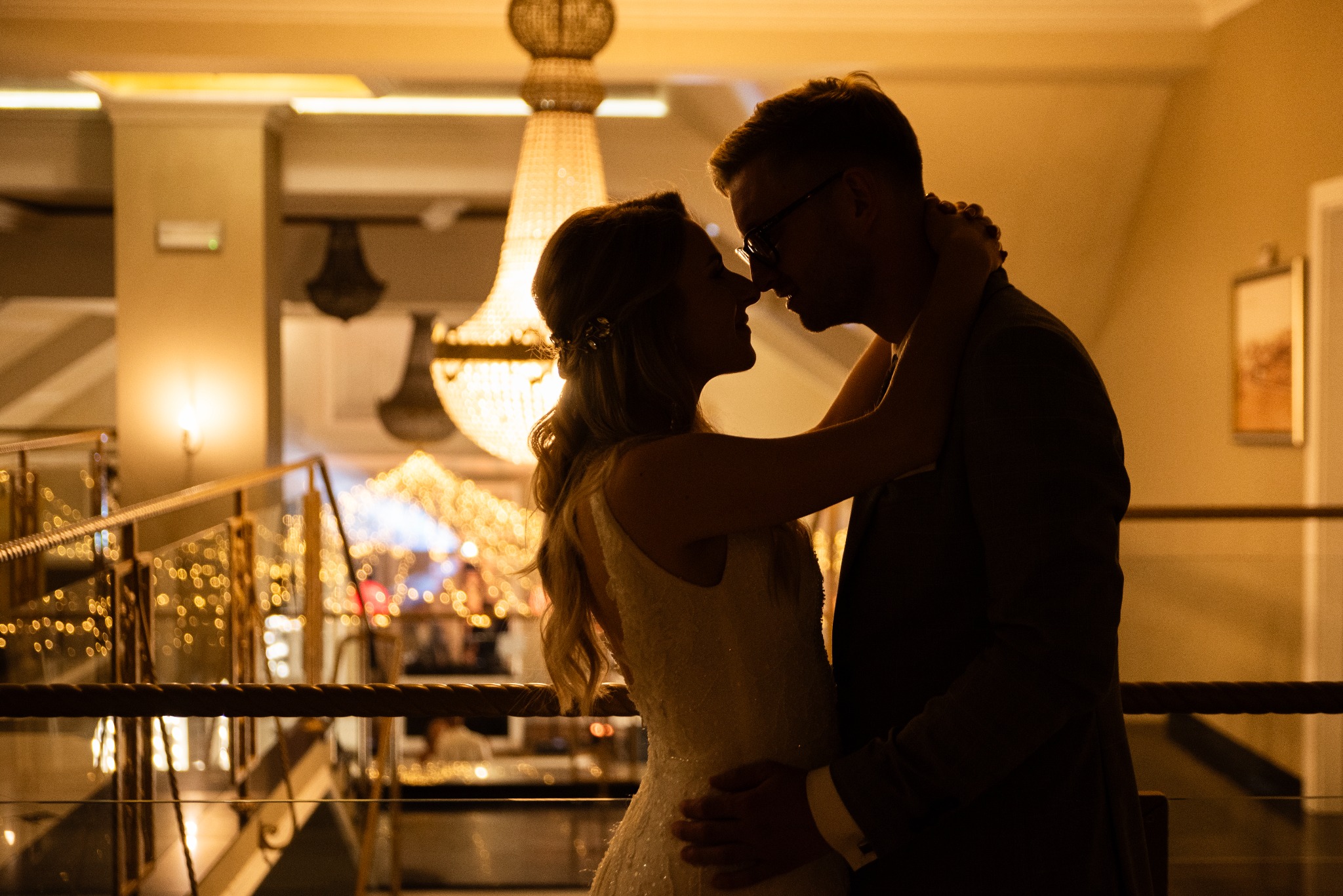 Silhouettes of a bride and groom embracing indoors, illuminated by a large chandelier, creating a romantic, warm atmosphere during their wedding reception.