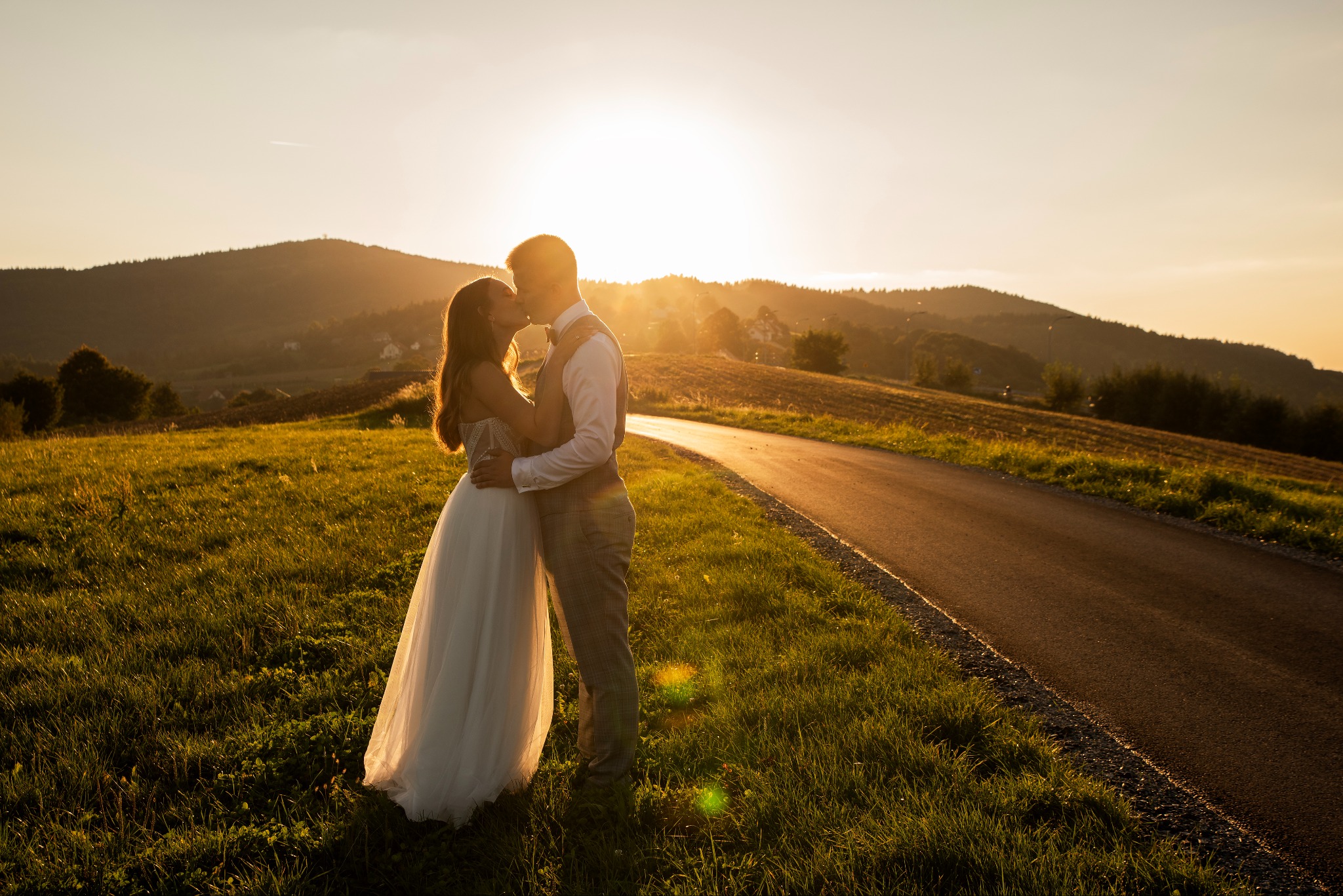 Para młoda w objęciach, pocałunek na tle zachodzącego słońca i malowniczych wzgórz. Romantyczna sesja ślubna w plenerze, ciepłe barwy zachodu.