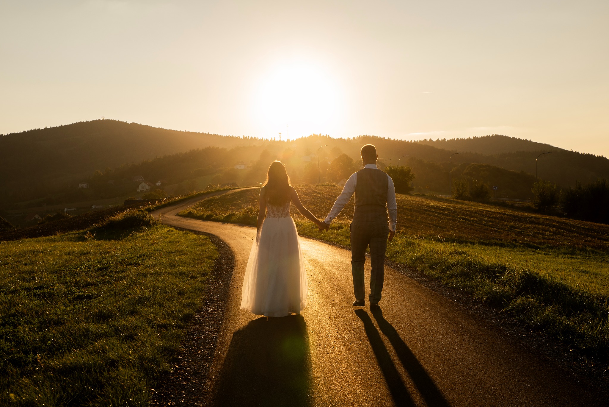 Para młoda, trzymając się za ręce, idzie drogą w kierunku zachodzącego słońca. W tle wzgórza i lasy, ciepłe, złote światło. Ujęcie z tyłu, podkreślające intymność chwili.