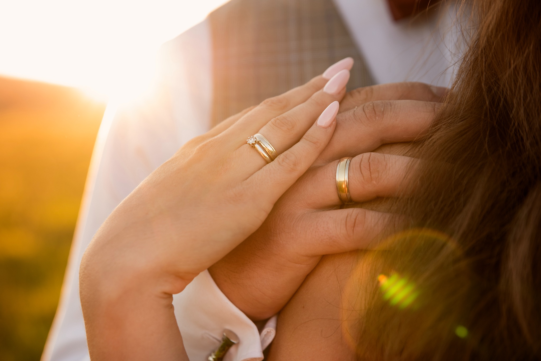 Zbliżenie dłoni pary młodej z złotymi obrączkami na tle rozświetlonego słońcem pleneru. Delikatne paznokcie kobiety i elegancki mankiet koszuli mężczyzny.