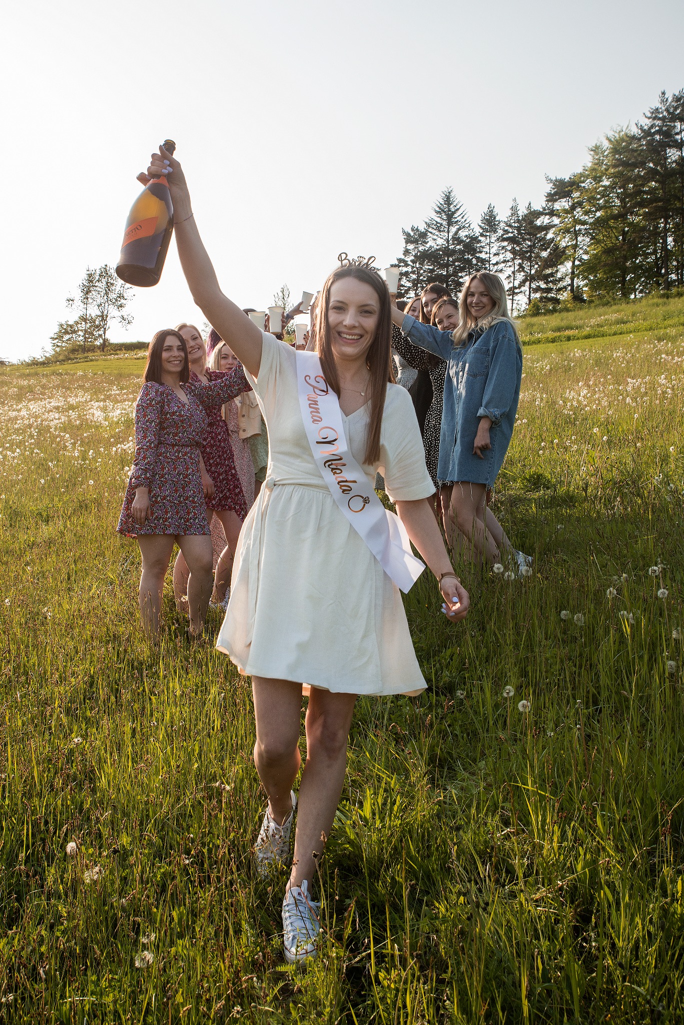 Uśmiechnięta przyszła panna młoda z szarfą i butelką szampana na łące, w tle druhny z plastikowymi kubkami. Słoneczny dzień, radosna atmosfera.