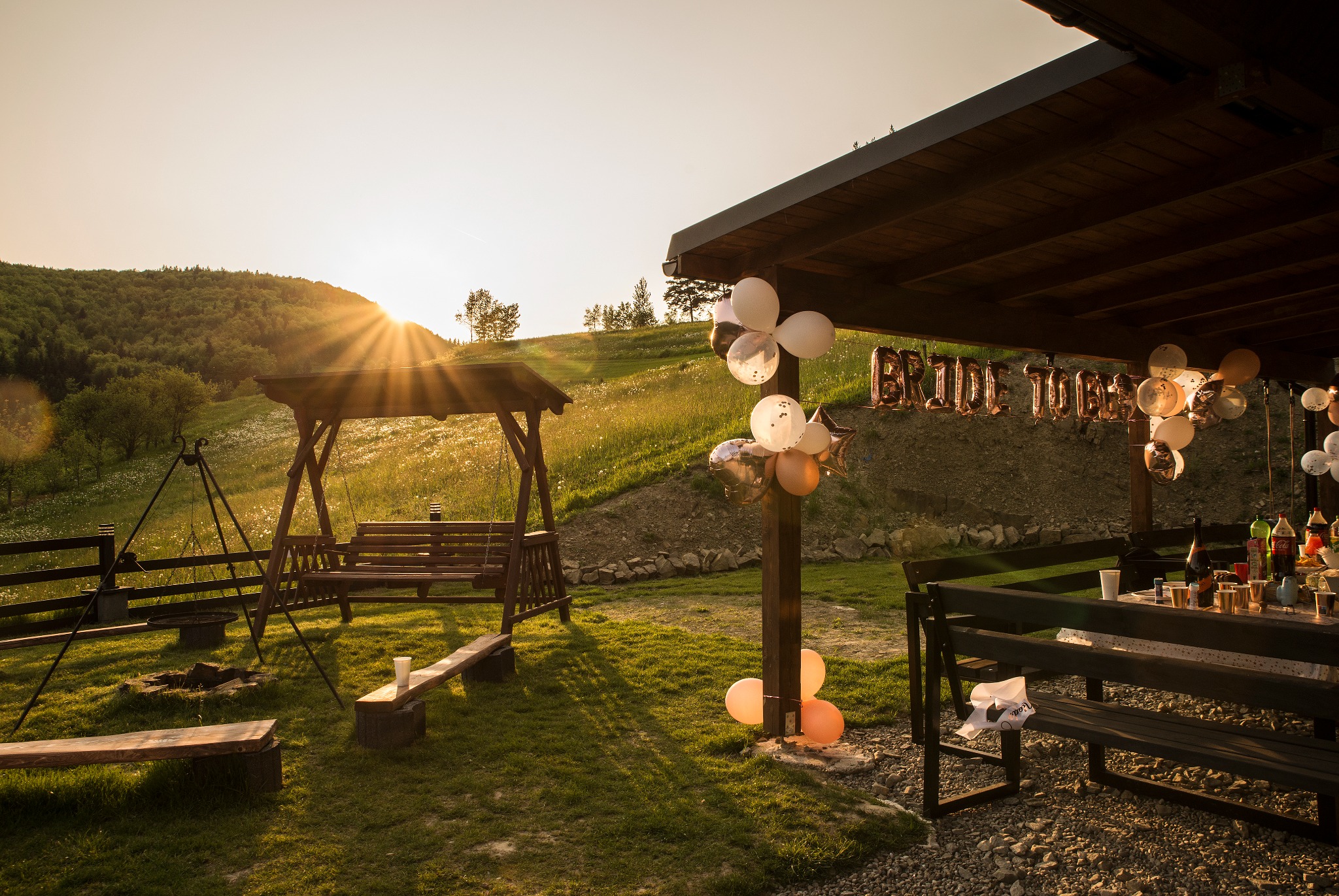 Plenerowa impreza 'Bride to be' w Brzesku o zachodzie słońca. Drewniana huśtawka, balony i stół z napojami na tle zielonych wzgórz.