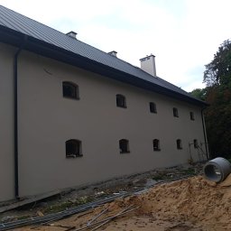 Adrian Orczyk - Świeżo otynkowany budynek z regularnie rozmieszczonymi oknami w kształcie łuku, widoczne rury kanalizacyjne i piasek przed budynkiem, sugerujące trwające prace wykończeniowe.
