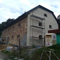 Adrian Orczyk - Kamienny budynek częściowo otynkowany, z rusztowaniem i stertą worków z zaprawą murarską, w tle widoczny las i pojemnik PCK.