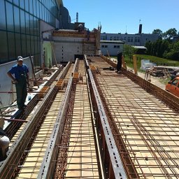 Adrian Orczyk - Przygotowanie szalunku i zbrojenia pod wylewkę stropu na budowie, widoczni robotnicy i pomarańczowy samochód dostawczy w tle.