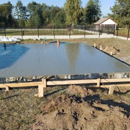 Adrian Orczyk - Wylana, świeża płyta fundamentowa pod budowę domu z widocznymi rurami instalacyjnymi, szalunkiem i czarną folią izolacyjną, na działce z trawnikiem i ogrodzeniem.