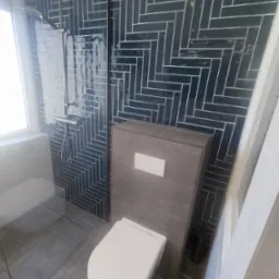 Nowoczesna łazienka z ciemnymi, błyszczącymi płytkami ułożonymi w jodełkę, szarą zabudową toalety podtynkowej i minimalistycznym prysznicem typu walk-in z odpływem liniowym.