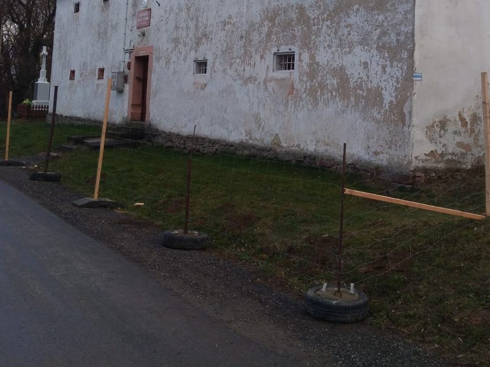 Stary budynek z uszkodzoną elewacją i prowizorycznym ogrodzeniem z siatki, wspartej na oponach i drewnianych słupkach.