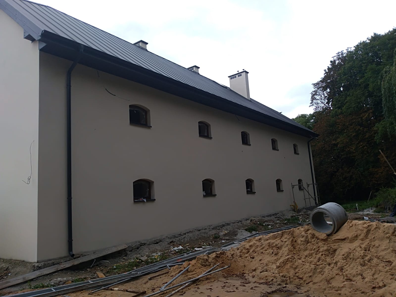 Świeżo otynkowany budynek z regularnie rozmieszczonymi oknami w kształcie łuku, widoczne rury kanalizacyjne i piasek przed budynkiem, sugerujące trwające prace wykończeniowe.