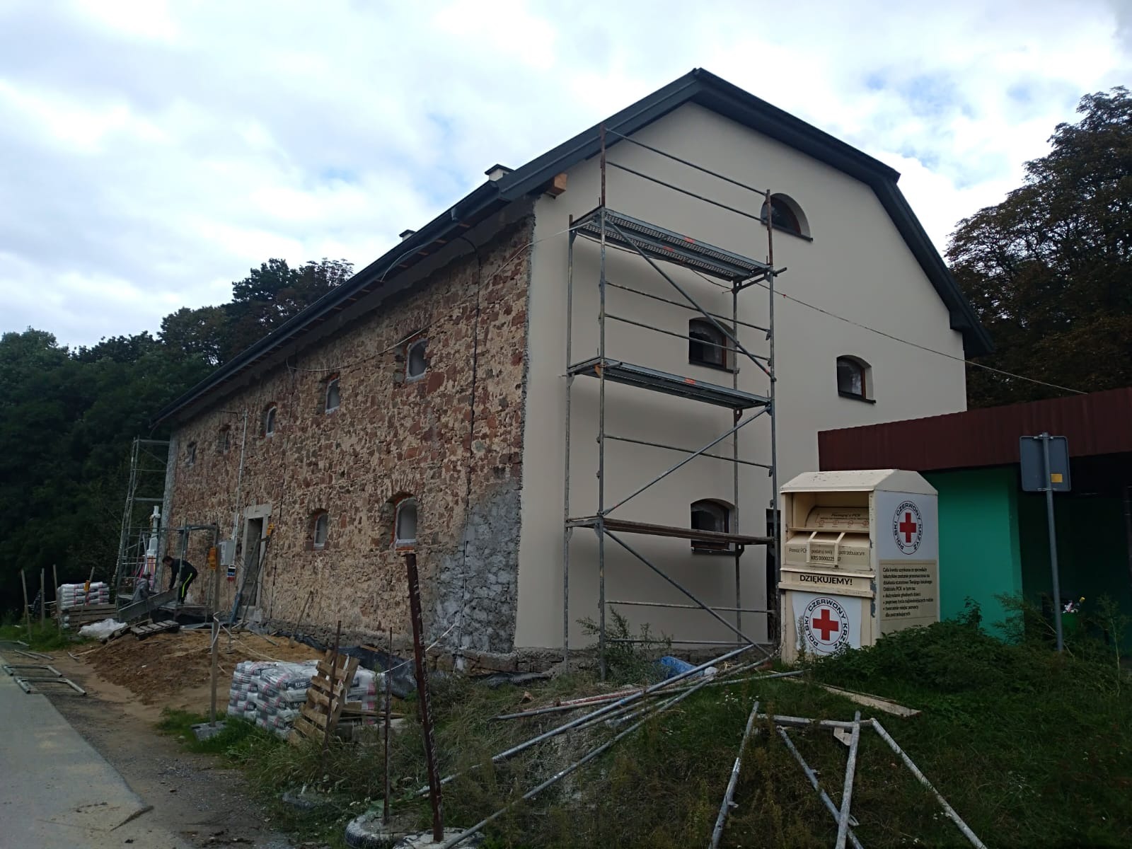Kamienny budynek częściowo otynkowany, z rusztowaniem i stertą worków z zaprawą murarską, w tle widoczny las i pojemnik PCK.