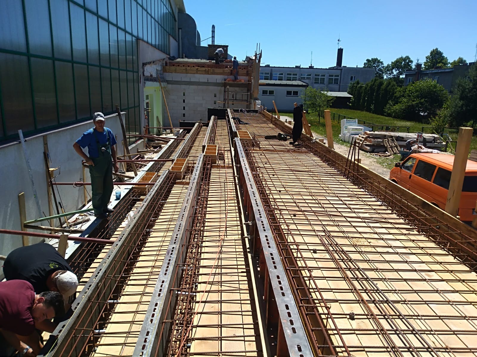 Przygotowanie szalunku i zbrojenia pod wylewkę stropu na budowie, widoczni robotnicy i pomarańczowy samochód dostawczy w tle.
