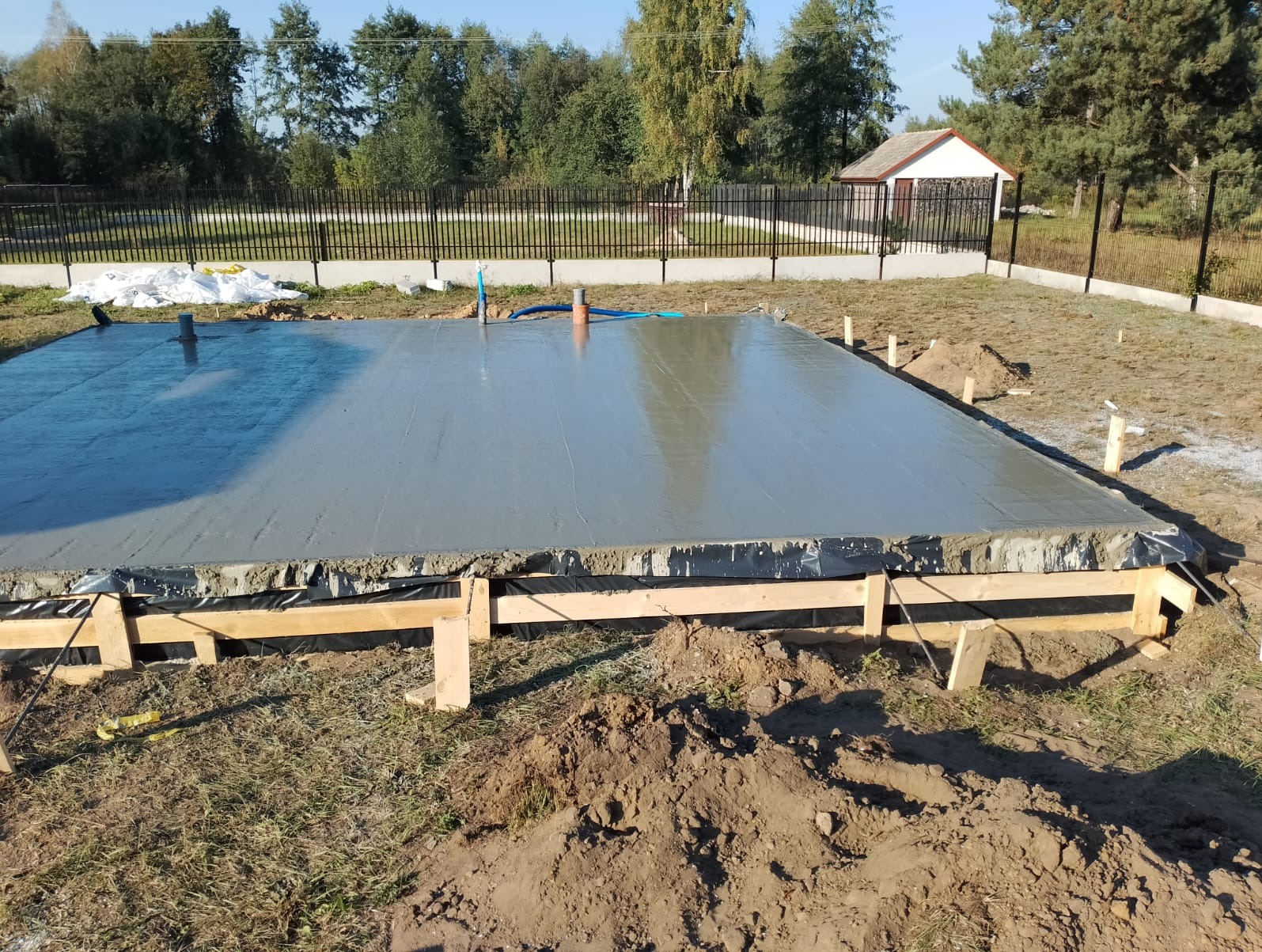 Wylana, świeża płyta fundamentowa pod budowę domu z widocznymi rurami instalacyjnymi, szalunkiem i czarną folią izolacyjną, na działce z trawnikiem i ogrodzeniem.
