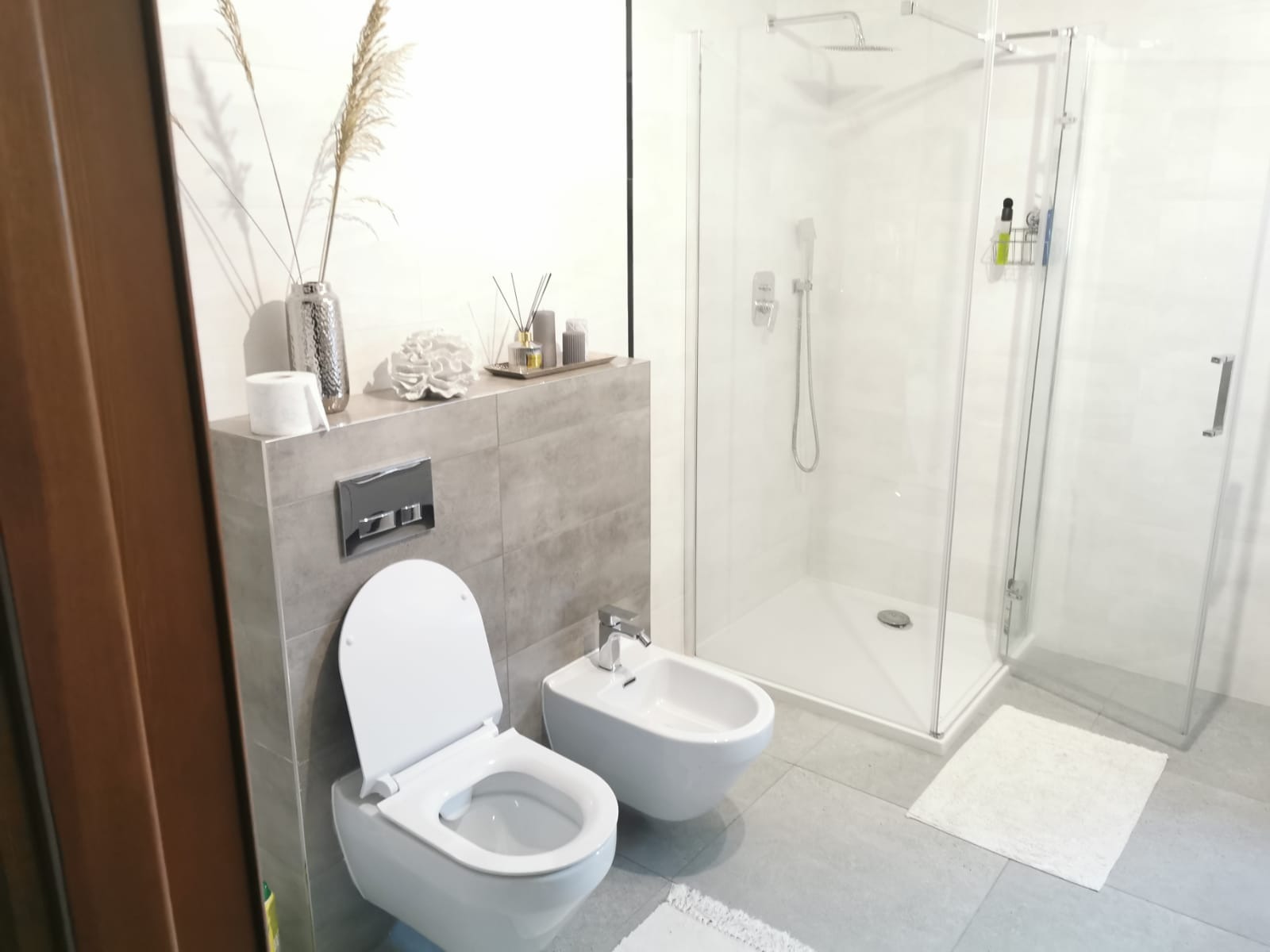 Nowoczesna łazienka w jasnych odcieniach szarości z białą armaturą, kabiną prysznicową typu walk-in, wiszącą toaletą i bidetem, udekorowana wazonem z suszonymi trawami i dyfuzorem zapachowym.