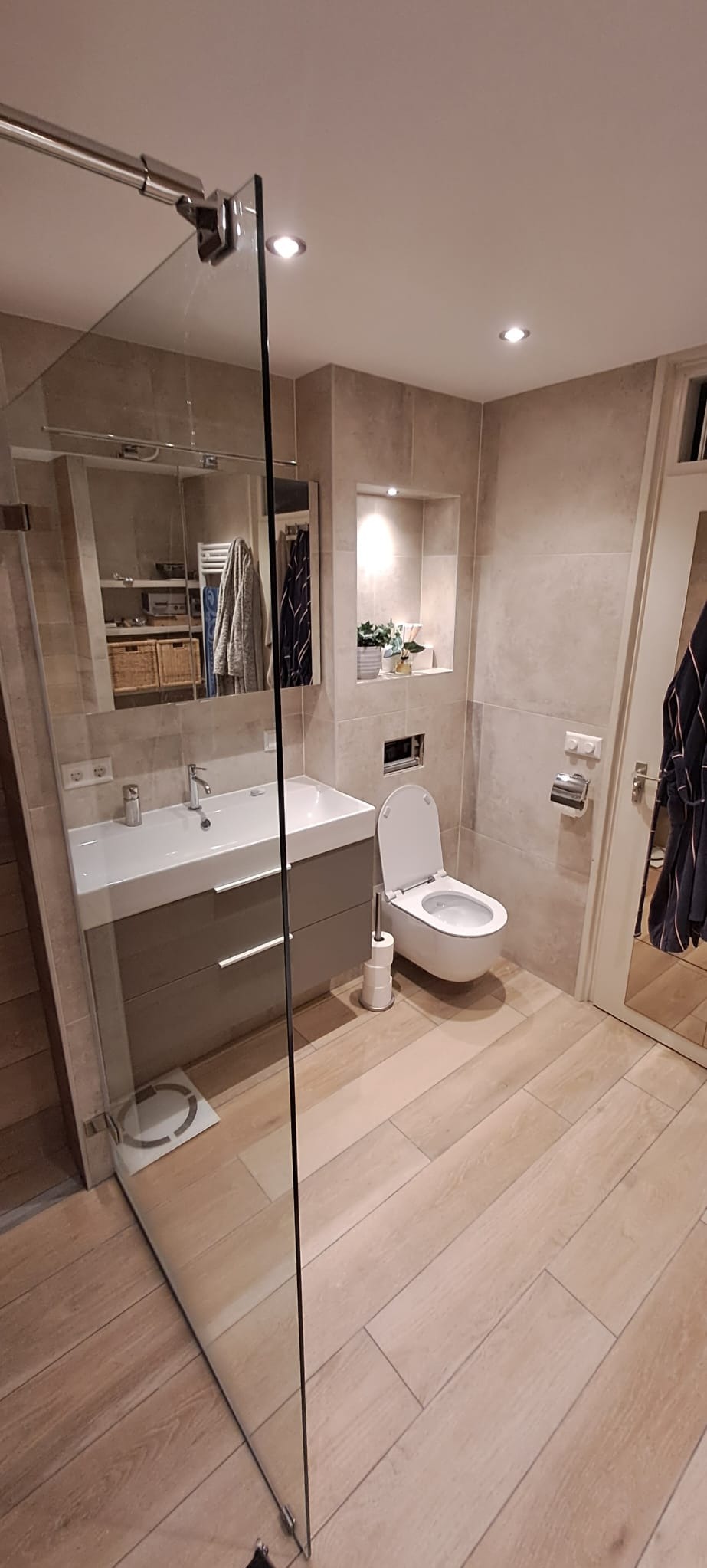 Jasna łazienka z drewnopodobną podłogą, szklaną kabiną prysznicową, umywalką z szufladami, toaletą i dekoracyjną wnęką z roślinami.