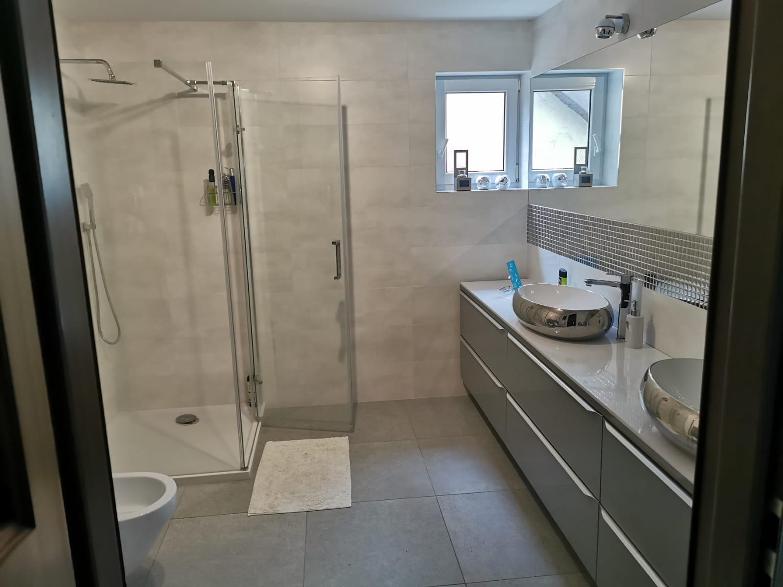 Nowoczesna łazienka z kabiną prysznicową typu walk-in, podwójną umywalką nablatową w kolorze srebrnym, szarą szafką z szufladami i mozaiką nad blatem.