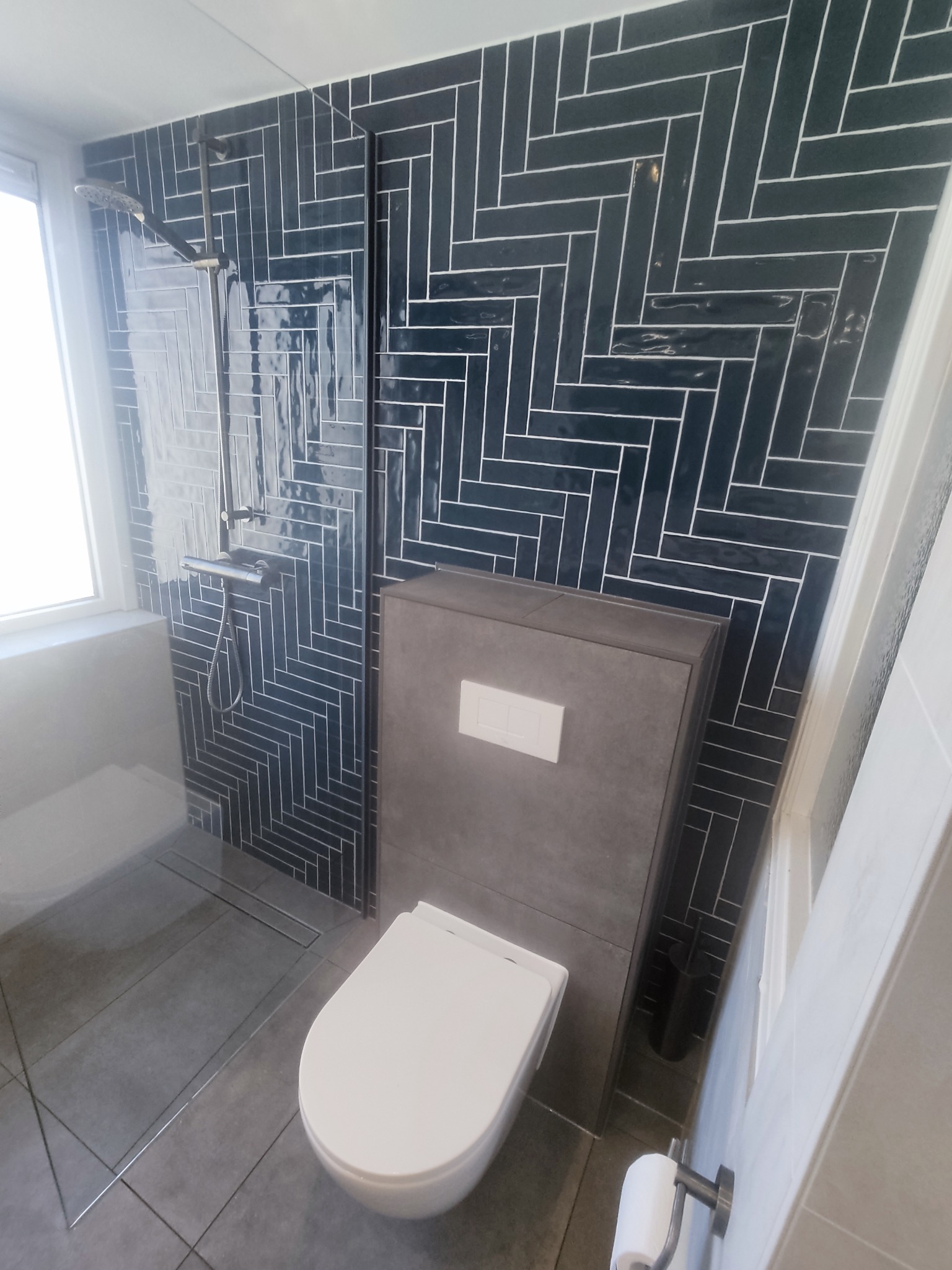 Nowoczesna łazienka z ciemnymi, błyszczącymi płytkami ułożonymi w jodełkę, szarą zabudową toalety podtynkowej i minimalistycznym prysznicem typu walk-in z odpływem liniowym.