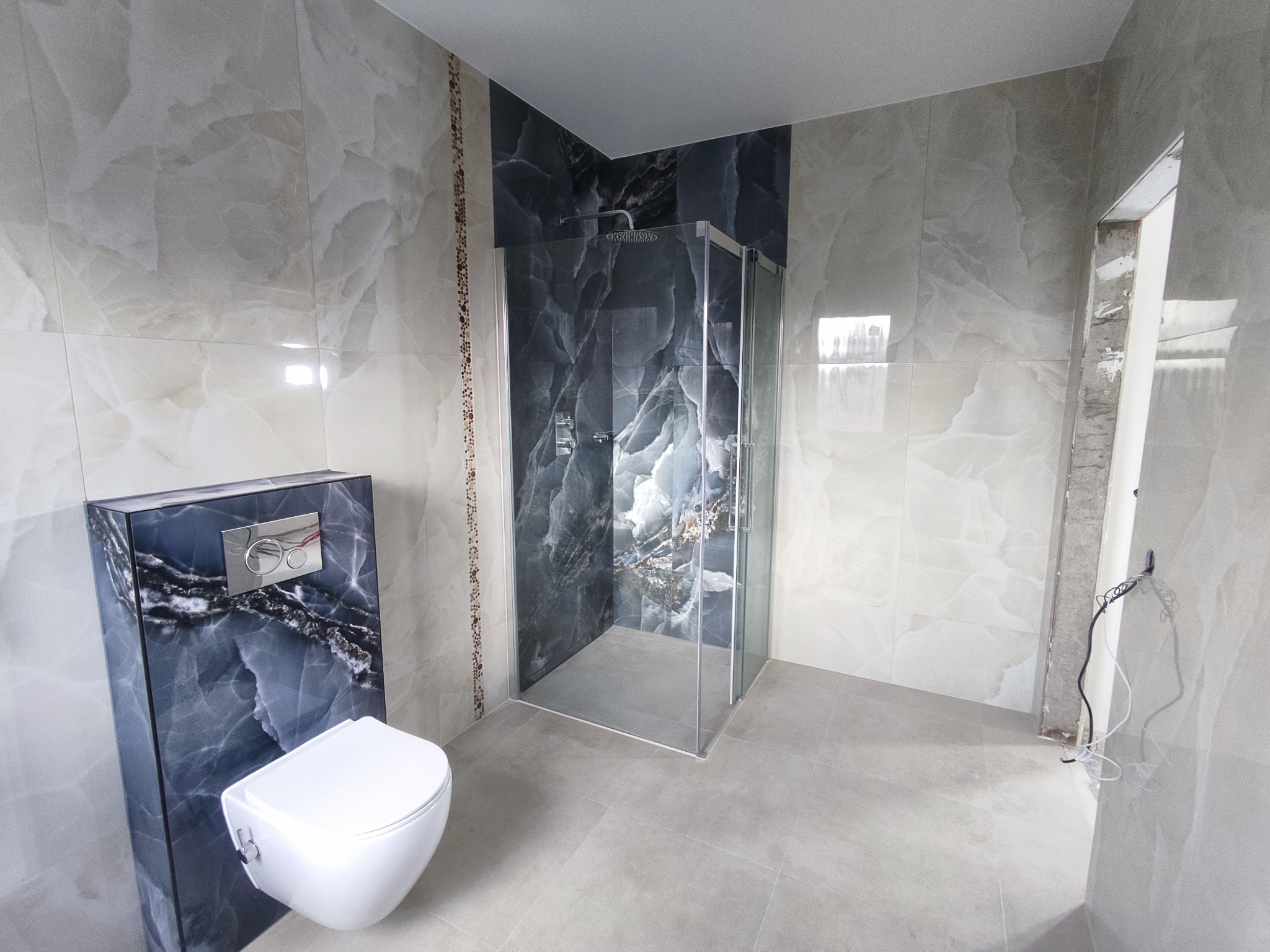 Nowoczesna łazienka z kabiną prysznicową typu walk-in, wiszącą toaletą i płytkami imitującymi marmur w odcieniach szarości i bieli, widoczne niedokończone prace elektryczne przy framudze drzwi.