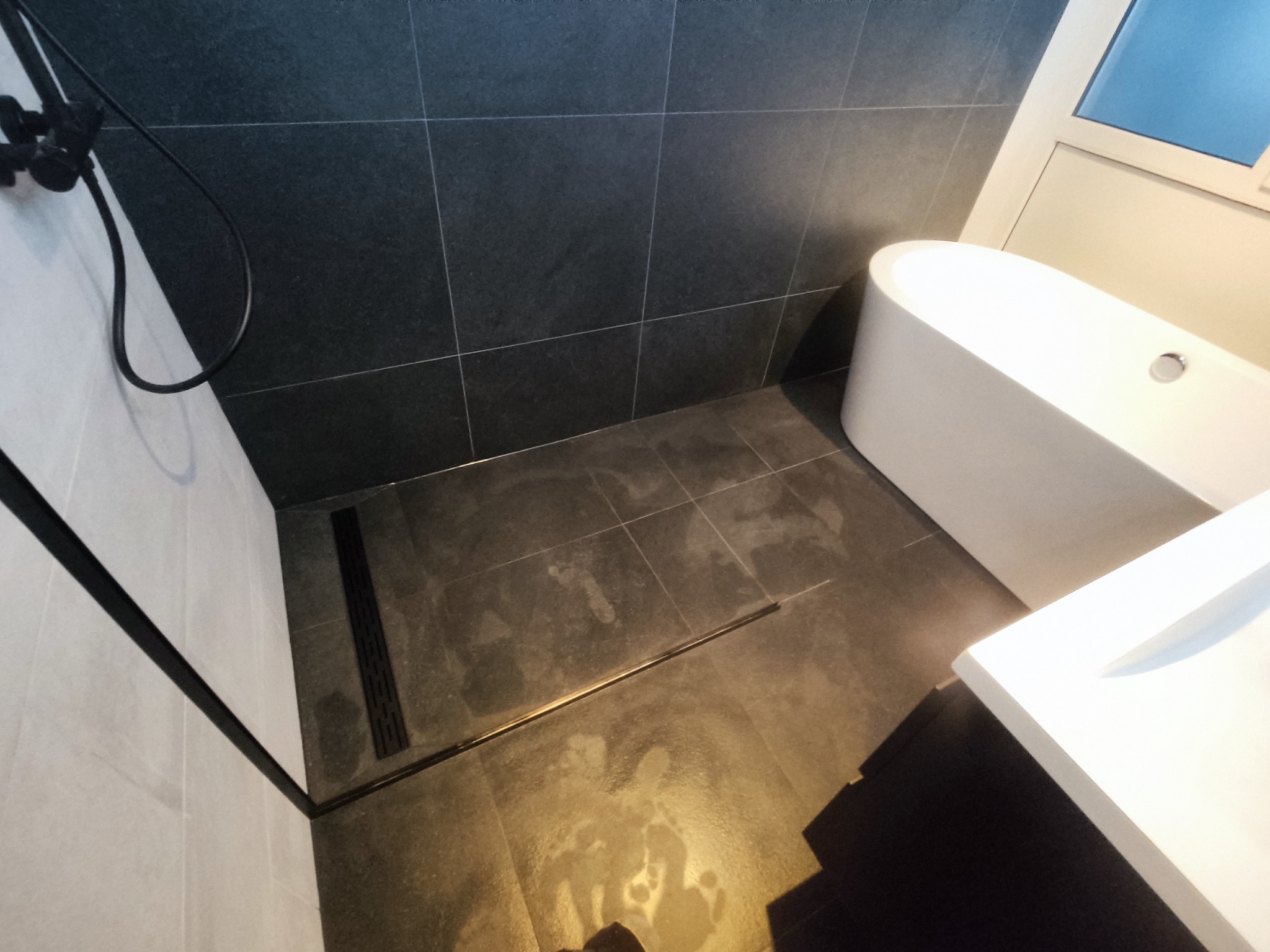 Nowoczesna łazienka z ciemnymi płytkami, minimalistycznym odpływem liniowym, białą wanną wolnostojącą i umywalką, widoczny fragment kabiny prysznicowej z czarną armaturą.