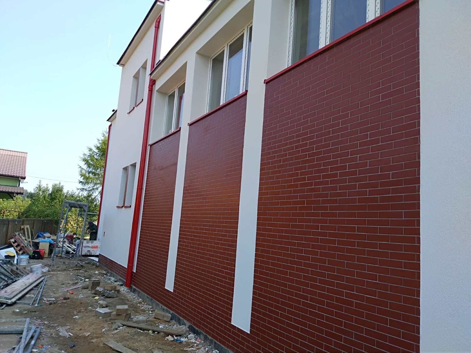 Dwukondygnacyjny budynek z częściowo wykończoną elewacją z brązowej cegły klinkierowej; widoczne rusztowanie i materiały budowlane.