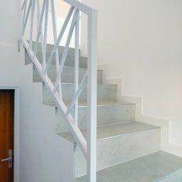 Michalski - Nowoczesne schody z szarymi stopniami i białą, geometryczną balustradą, widok z dołu, drewniane drzwi po lewej stronie.