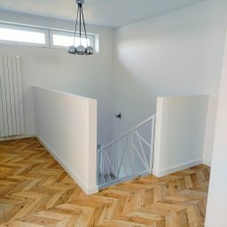 Michalski - Nowoczesne wnętrze z parkietem ułożonym w jodełkę, białymi ścianami i designerską lampą wiszącą z trzema chromowanymi kulami, widok na schody z białą balustradą.