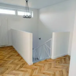 Nowoczesne wnętrze z parkietem ułożonym w jodełkę, białymi ścianami i designerską lampą wiszącą z trzema chromowanymi kulami, widok na schody z białą balustradą.