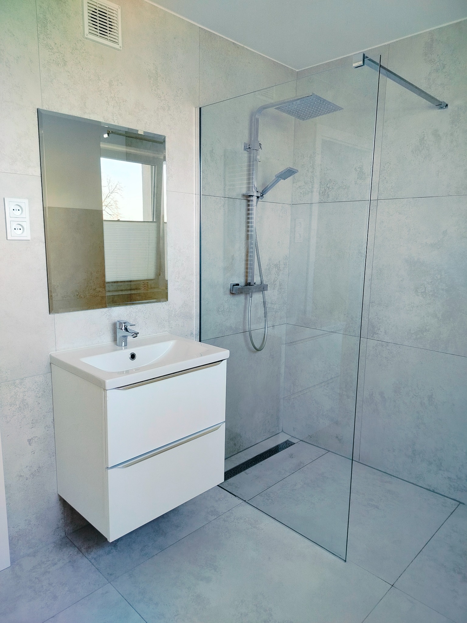 Nowoczesna łazienka z kabiną prysznicową typu walk-in, umywalką nablatową z białą szafką z dwoma szufladami, lustrem i szarą, teksturowaną płytką na ścianach i podłodze.