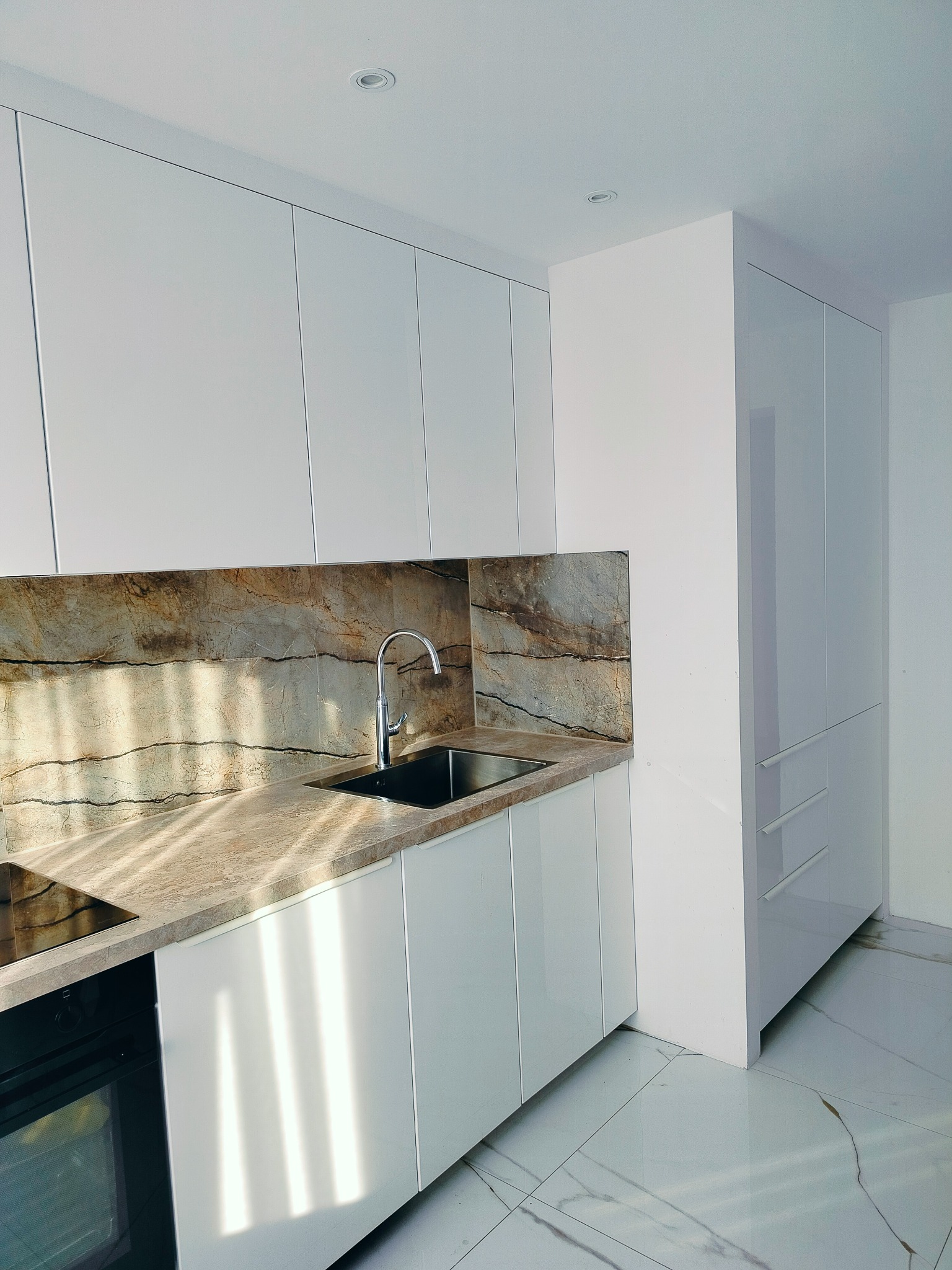 Nowoczesna kuchnia z białymi lakierowanymi szafkami, blatem i backsplashiem z imitacji marmuru oraz połyskującą podłogą z płytek gresowych.