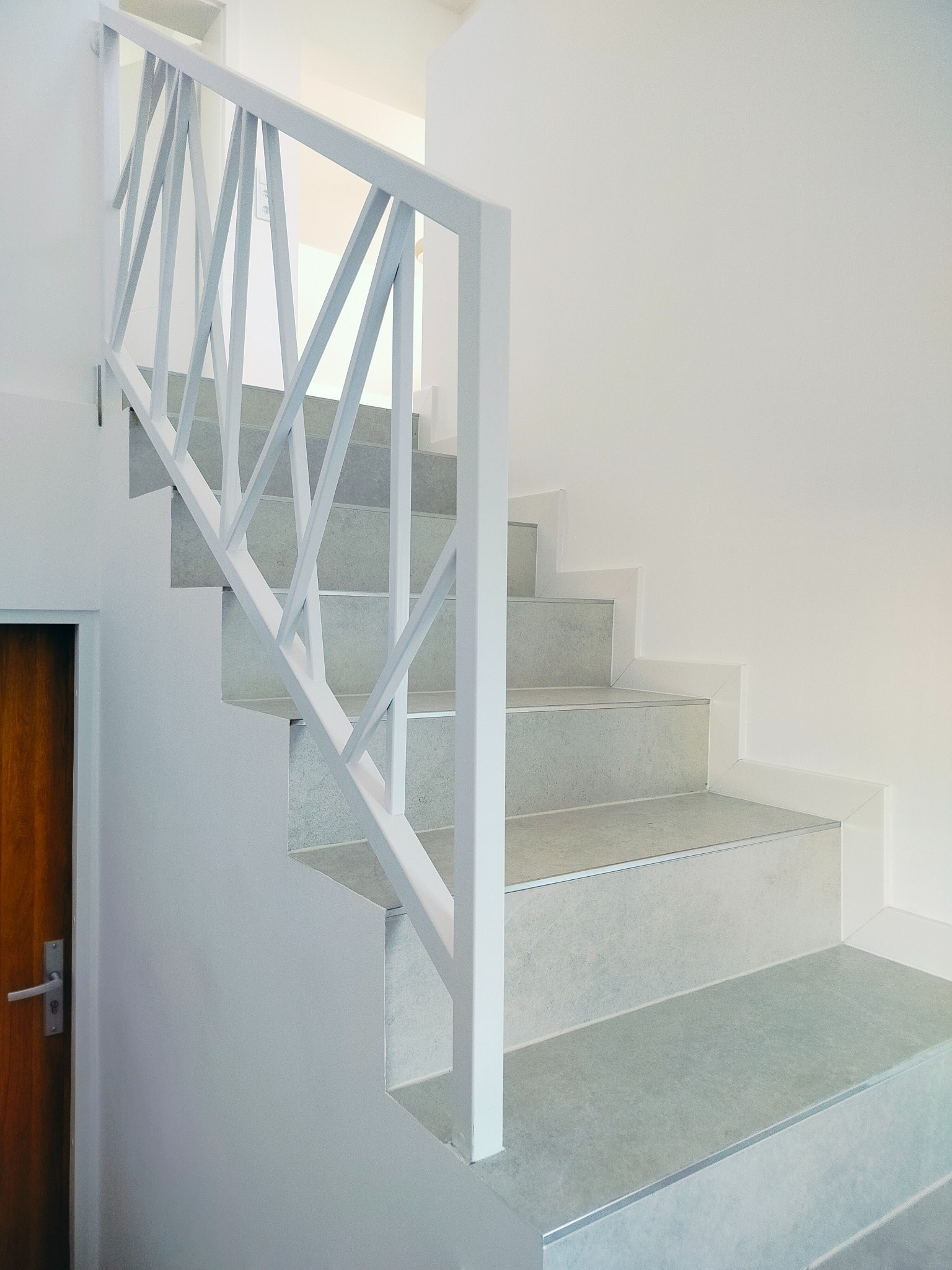 Nowoczesne schody z szarymi stopniami i białą, geometryczną balustradą, widok z dołu, drewniane drzwi po lewej stronie.