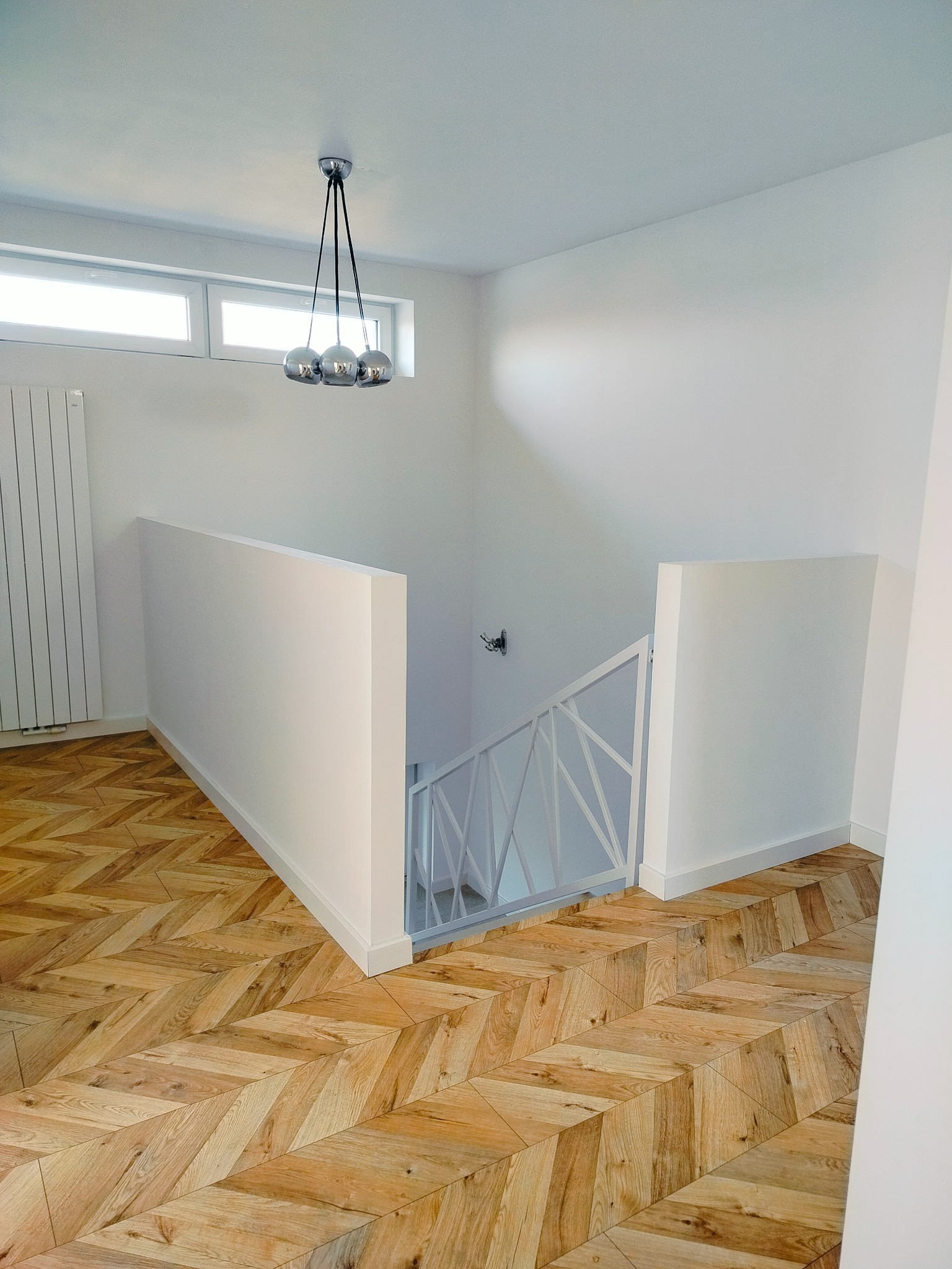 Nowoczesne wnętrze z parkietem ułożonym w jodełkę, białymi ścianami i designerską lampą wiszącą z trzema chromowanymi kulami, widok na schody z białą balustradą.