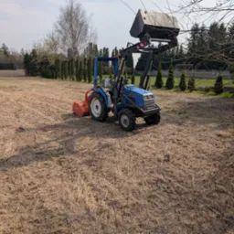 Niebieski traktor Iseki z osprzętem na polu z resztkami trawy, w tle rząd tuj i drzewa. Sprzęt gotowy do pracy.