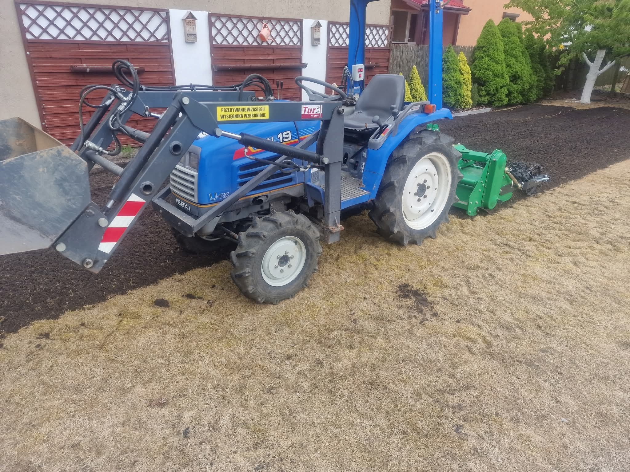 Niebieski traktor ISEKI z łyżką i glebogryzarką na trawniku, obok świeżo przekopana ziemia. Widoczne elementy ogrodzenia.