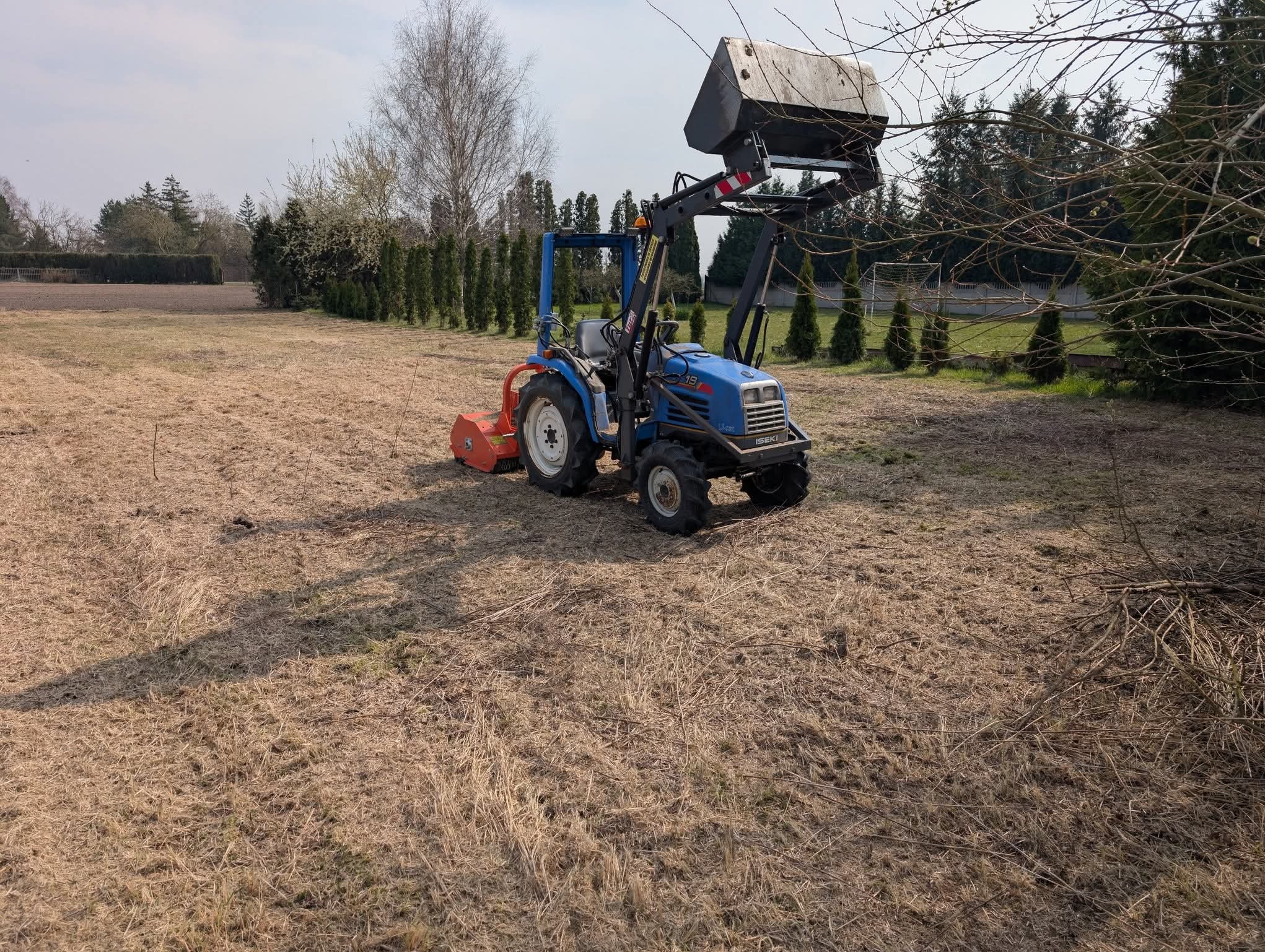 Niebieski traktor Iseki z osprzętem na polu z resztkami trawy, w tle rząd tuj i drzewa. Sprzęt gotowy do pracy.
