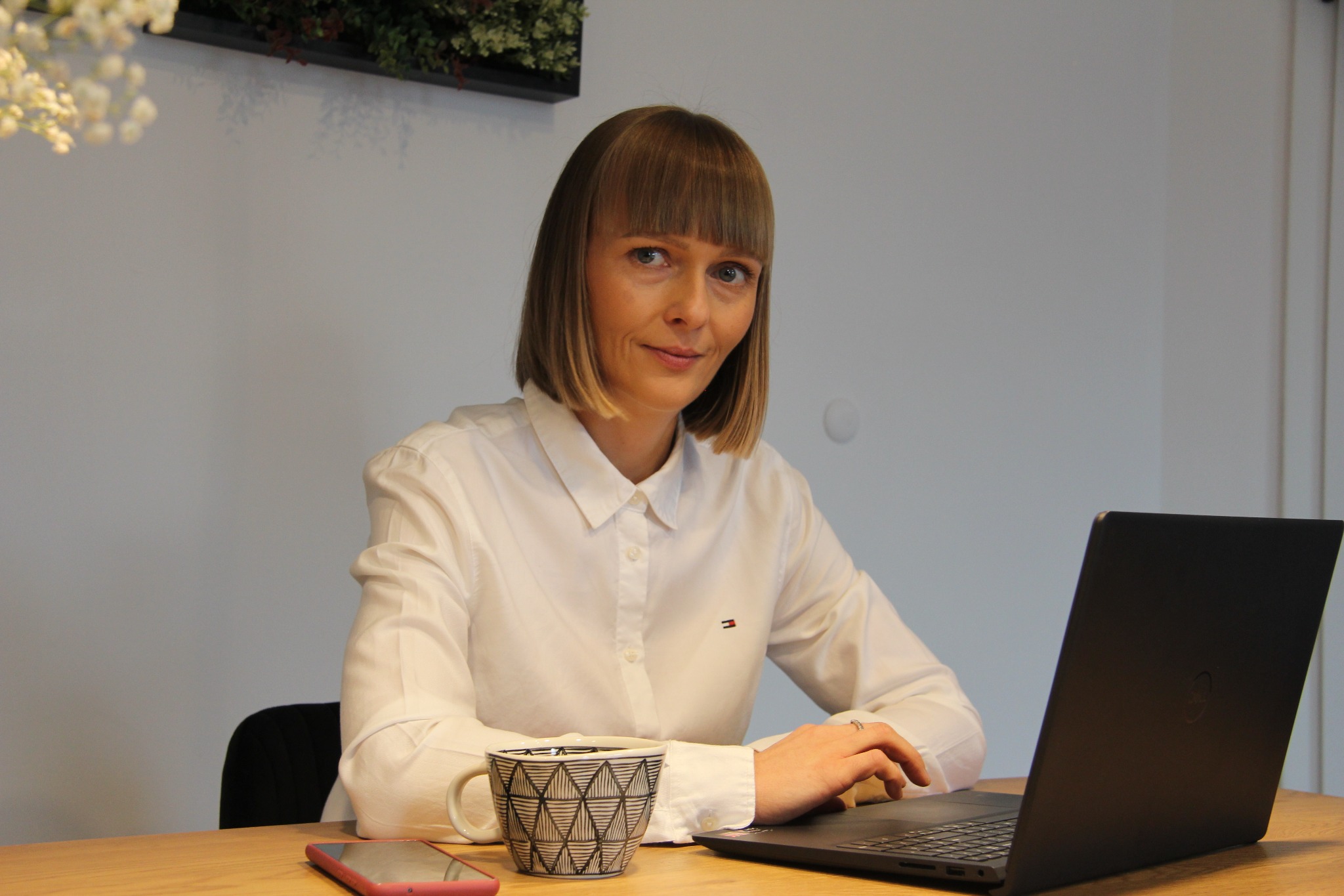 Kobieta z krótkimi blond włosami w białej koszuli, siedząca przy biurku z laptopem, telefonem i kubkiem z geometrycznym wzorem, uśmiechnięta w stronę aparatu.