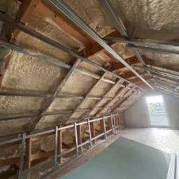 Poddasze z ociepleniem pianką PUR, widoczne drewniane belki i metalowy stelaż pod płyty gipsowe, okno w tle. Izolacja natryskowa na skośnym dachu.