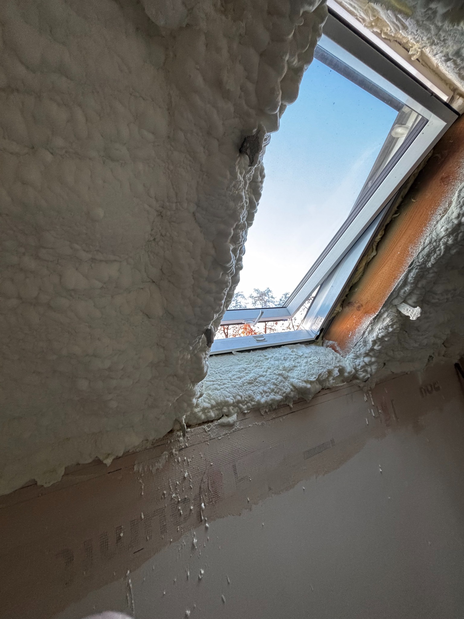 Poddasze z oknem połaciowym, pokryte grubą warstwą białej pianki izolacyjnej, widoczne niedoskonałości w nałożeniu i resztki pianki na ścianie, z widokiem na błękitne niebo i korony drzew przez...