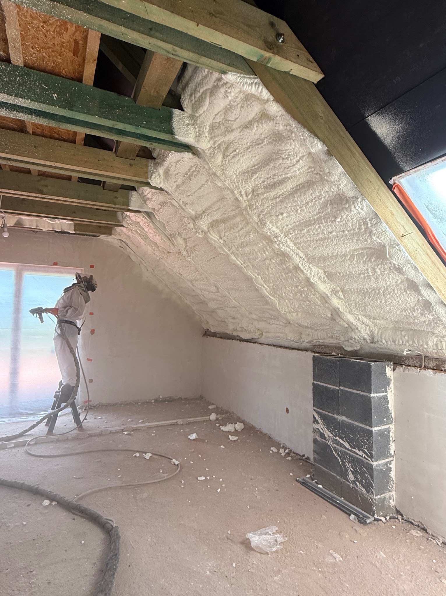 Pracownik w kombinezonie aplikuje piankę PUR na poddasze domu. Widoczne drewniane elementy konstrukcyjne dachu i świeżo nałożona izolacja.