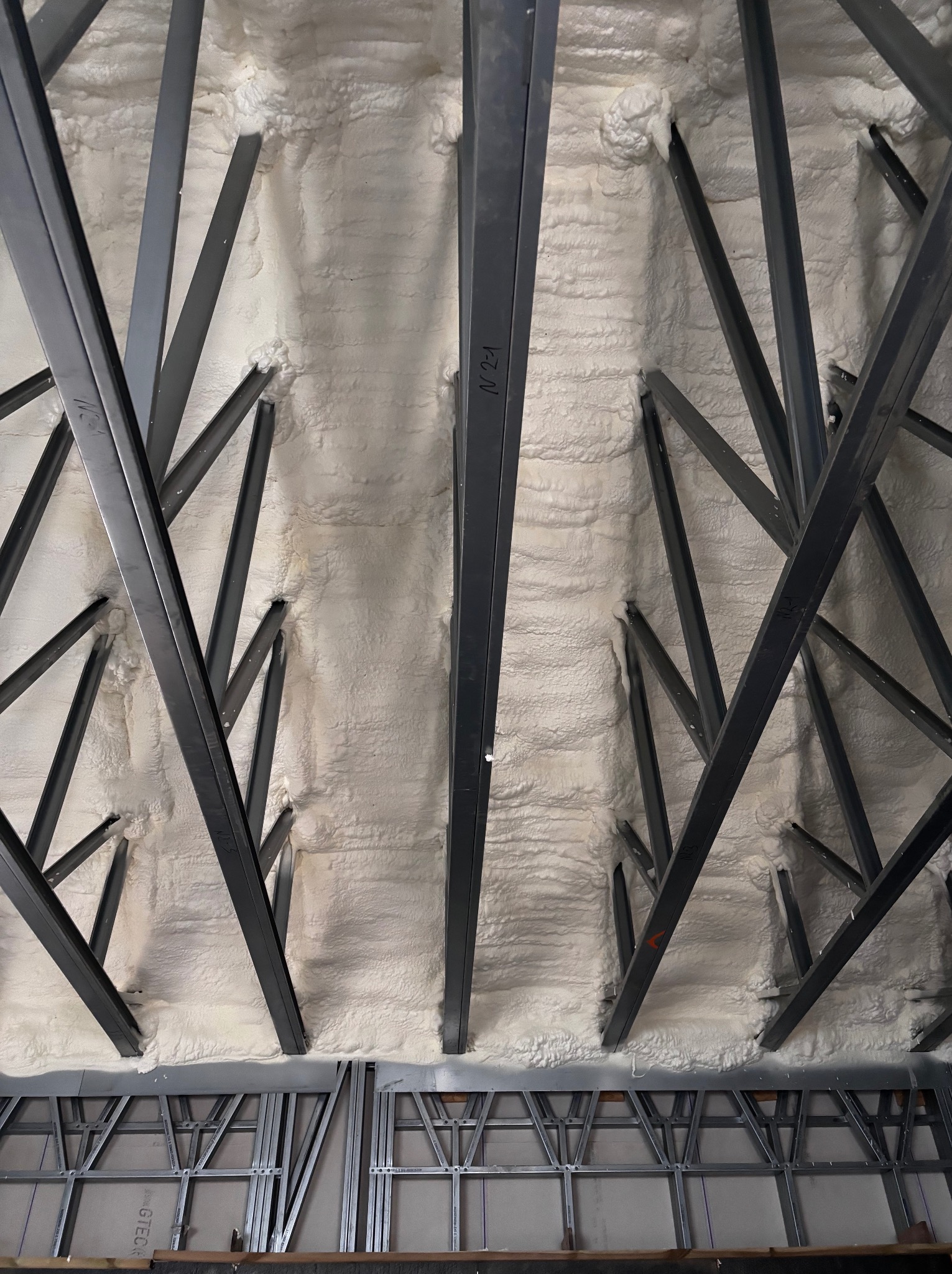 Poddasze z widoczną konstrukcją metalową i zaaplikowaną białą pianką izolacyjną PUR, wypełniającą przestrzeń między elementami konstrukcyjnymi.