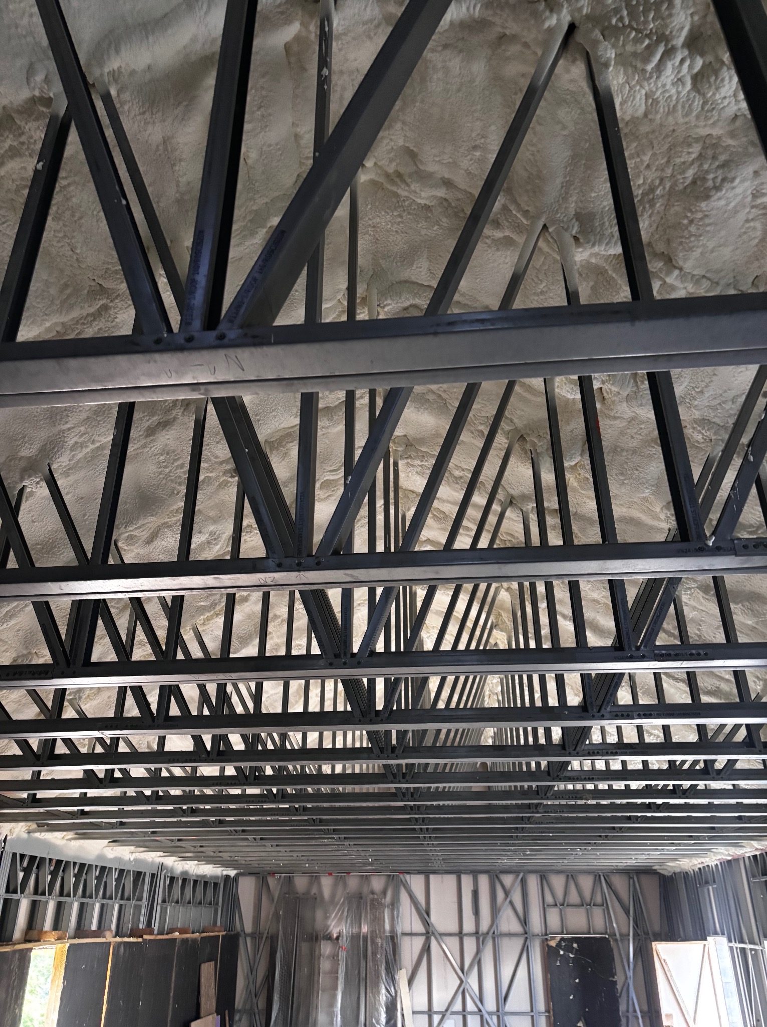 Poddasze z widoczną konstrukcją stalową i zaaplikowaną pianką PUR, tworzącą jednolitą warstwę izolacyjną. Widok z dołu na ocieplony strop.