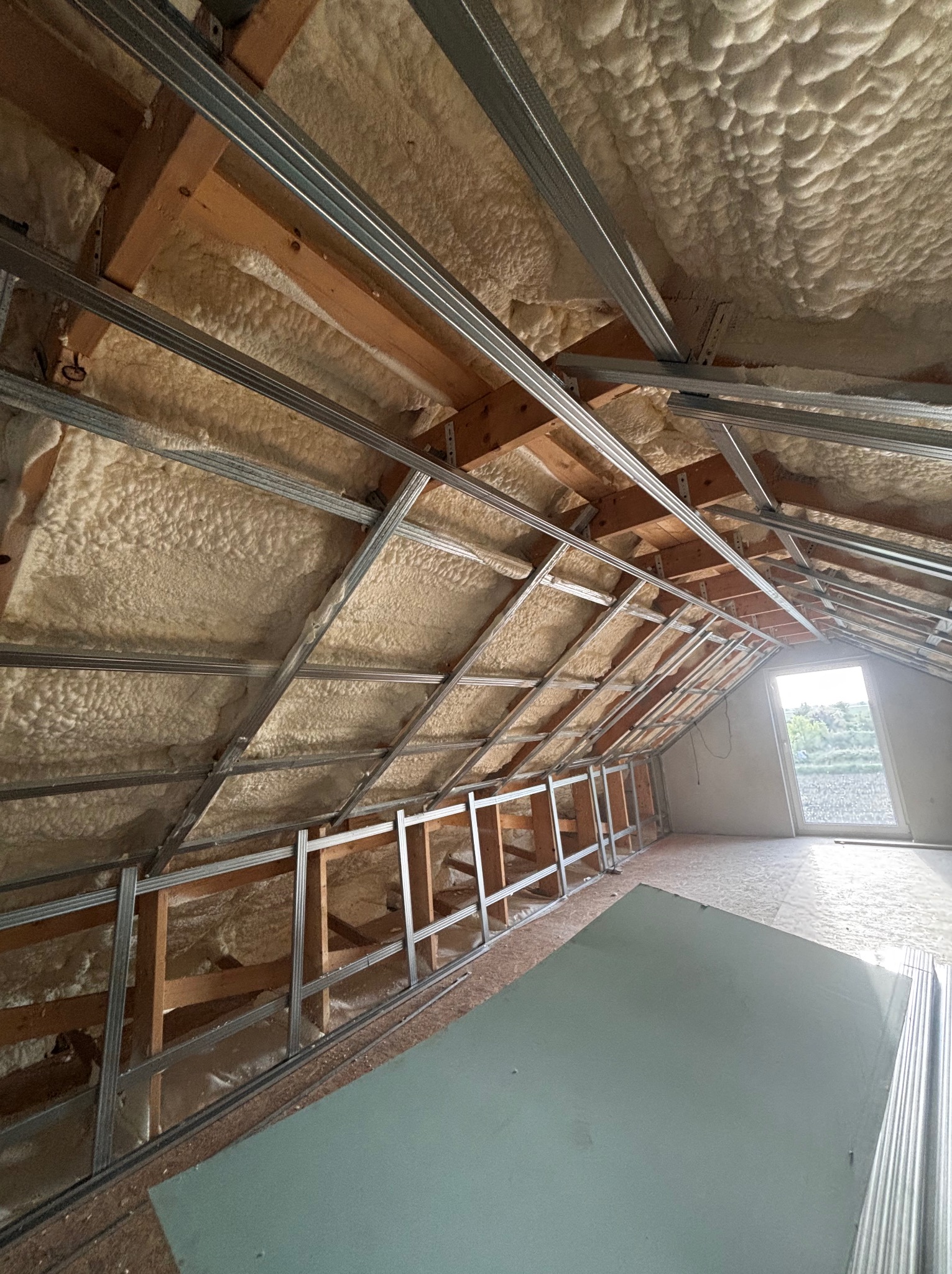 Poddasze z ociepleniem pianką PUR, widoczne drewniane belki i metalowy stelaż pod płyty gipsowe, okno w tle. Izolacja natryskowa na skośnym dachu.
